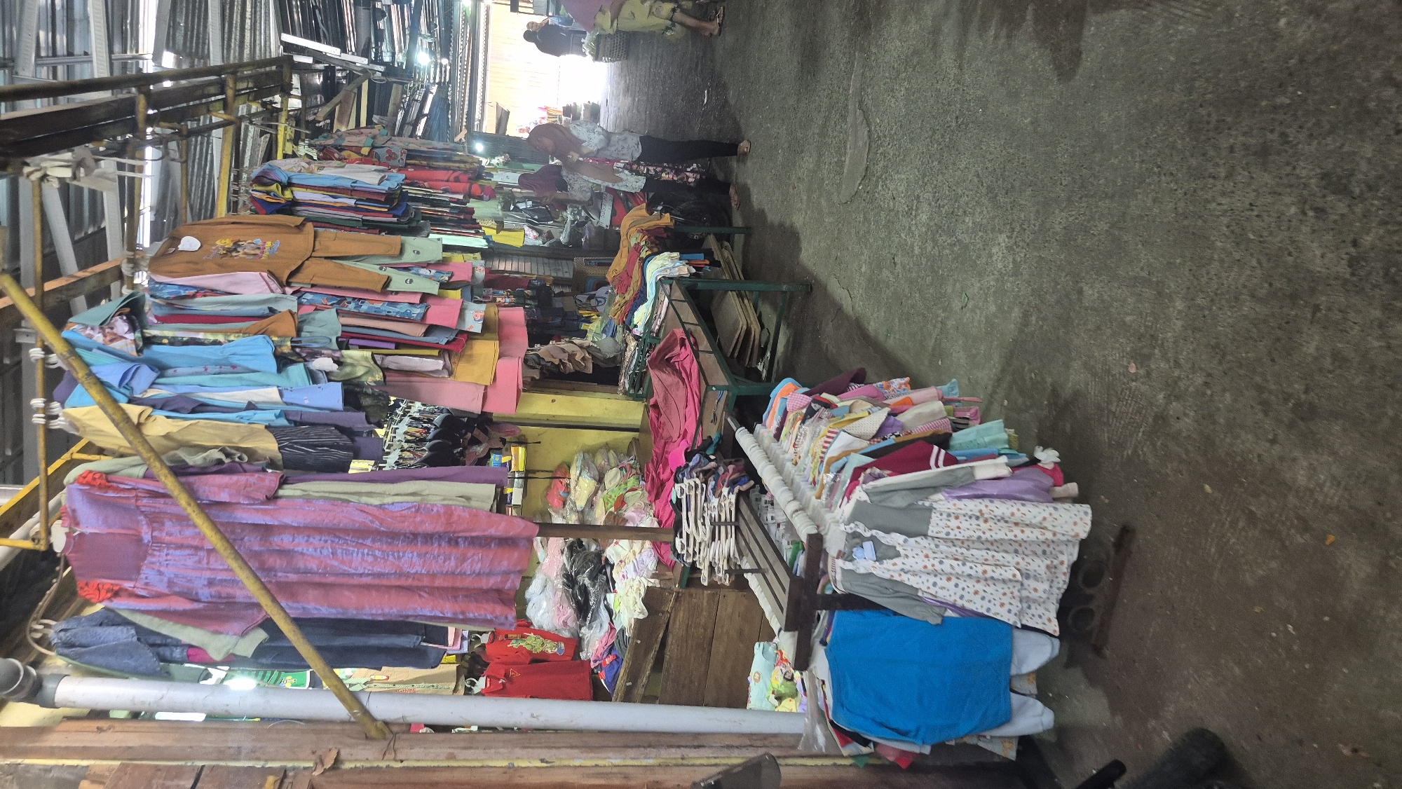 The image depicts a clothing market stall in Boja, Indonesia, with a variety of garments displayed on racks and tables. The stall features an assortment of apparel, including dresses, shirts, and pants, predominantly in bright colors and varied patterns. Some clothing items are neatly hung on hangers, while others are piled on surfaces. The environment appears to be an open-air market with a corrugated metal roof visible in the background, suggesting a covered yet exposed setting. Two individuals are visible in the mid-ground, appearing to be shoppers or vendors, one of whom is wearing a hijab and a patterned outfit, while the other is partially obscured. The ground is paved with concrete, and there are wooden structures and metal racks supporting the merchandise. The overall scene captures the bustling atmosphere of a local market.