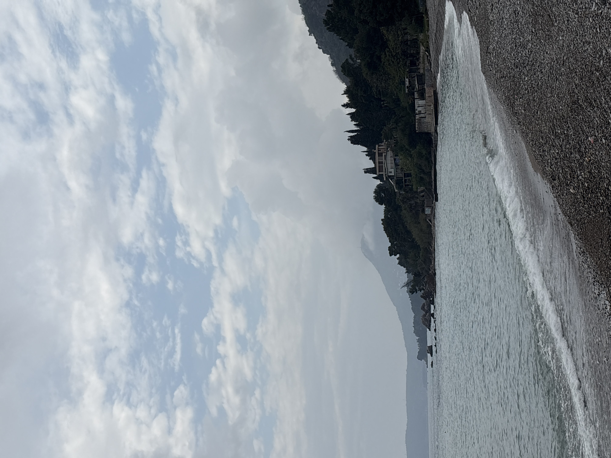 The image displays a tranquil coastal landscape, likely in Bar, Montenegro, featuring a pebble beach, the sea, and a mountainous shoreline under a cloudy sky.
The sky, occupying the upper portion, is mostly covered by light grey and white clouds, with patches of pale blue visible between them.
To the left, a steep, forested mountain range rises, densely covered in green trees. Several light-colored buildings with red roofs are visible nestled among the trees along the lower slopes, suggesting a coastal settlement. Further in the distance, additional layers of mountains appear hazy and lighter, fading into the horizon.
The sea, a light blue-grey hue, fills the middle ground, appearing relatively calm. Gentle white waves are consistently breaking along the shore, creating foamy lines where they meet the land.
The foreground and extending to the right is a wide pebble beach, composed of small, dark grey and brown stones. No people or specific activities are visible, contributing to a serene and natural atmosphere. The diffused lighting suggests it is daytime, possibly an overcast day. There is no visible text in the image.