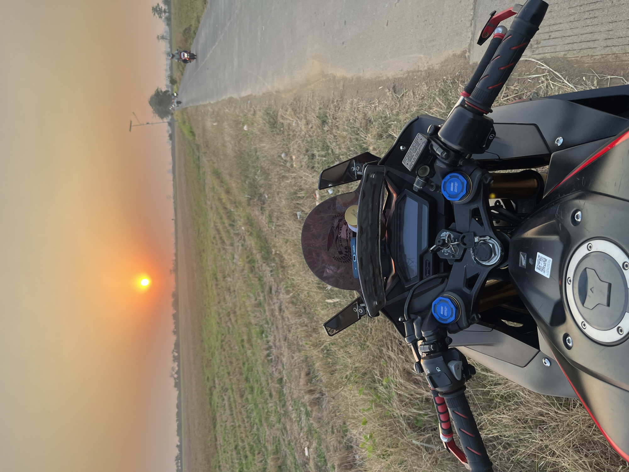 The image shows a first-person view from a motorcycle parked on the side of a paved road. The road is bordered by dry grass and fields. In the background, the sun is setting, casting a warm orange glow over the scene. Several other motorcycles are visible on the road in the distance. The motorcycle in the foreground is black with red accents and has a digital display on the dashboard. The location is identified as Troso, Indonesia.
