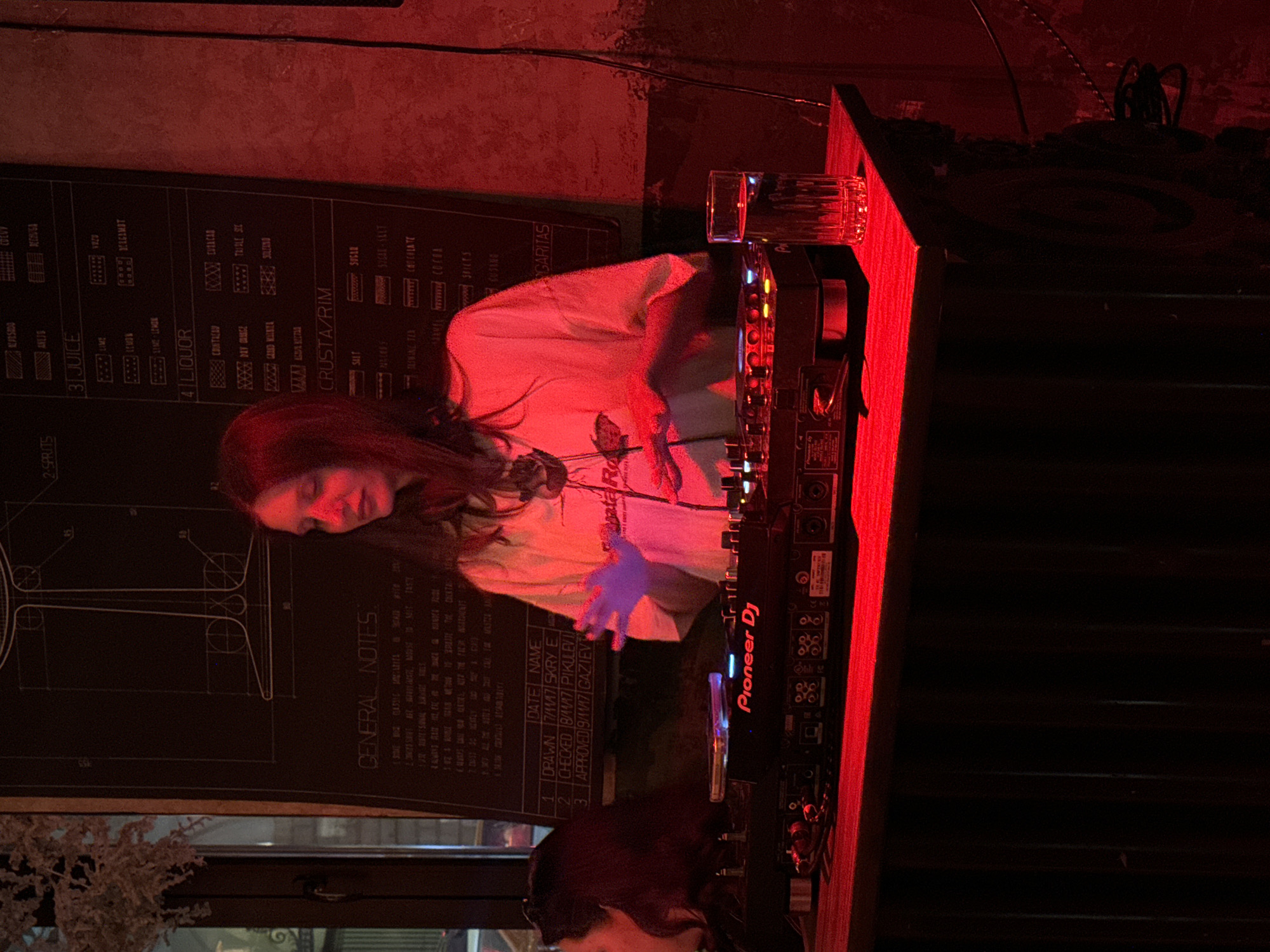 The image displays an indoor scene, characterized by strong red ambient lighting, in what appears to be a bar or club setting within Moscow, Russia. The central subject is a female individual with long, dark hair, seen from the waist up. She wears a light-toned, short-sleeved t-shirt with a dark graphic on the chest. Her hands are elevated above and interacting with a black Pioneer DJ mixer/controller, which is positioned on a dark wooden counter. The mixer's 'Pioneer DJ' logo is illuminated in red. To the immediate left of the central subject, a partial profile of another individual with dark hair is visible. A clear, ribbed glass containing liquid is situated on the counter to the right of the DJ equipment. The background features a large, dark rectangular board displaying white text and diagrams, including sections labeled 'GENERAL NOTES,' 'JUICE,' 'LIQUOR,' and 'CRUSTA/RIM,' along with dates and names such as 'SKRY E,' 'PIKULEV,' and 'GAZIEV.' On the far left, a blurred view of an exterior or window is present, alongside a dried floral or plant arrangement suspended overhead. The scene includes visible cables extending downwards along the right wall.