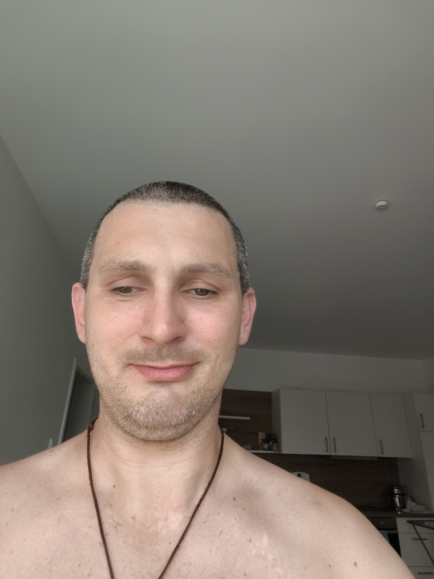 A man, likely in his late 20s to early 30s, is photographed from the chest up, in what appears to be an indoor setting. He has short, dark hair and a light beard. His head is tilted slightly, and he is looking downwards with a subtle smile. He is shirtless, wearing a thin, dark cord necklace. The background shows a plain ceiling and what looks like a kitchen area with white cabinets and a wooden backsplash. There is a small, white, circular object mounted on the ceiling, possibly a smoke detector. The lighting suggests it is daytime, with natural light coming from the left side of the frame. The man is positioned in the foreground, with the background blurred. The image does not provide any specific clues about the location within Wolgast, Germany.