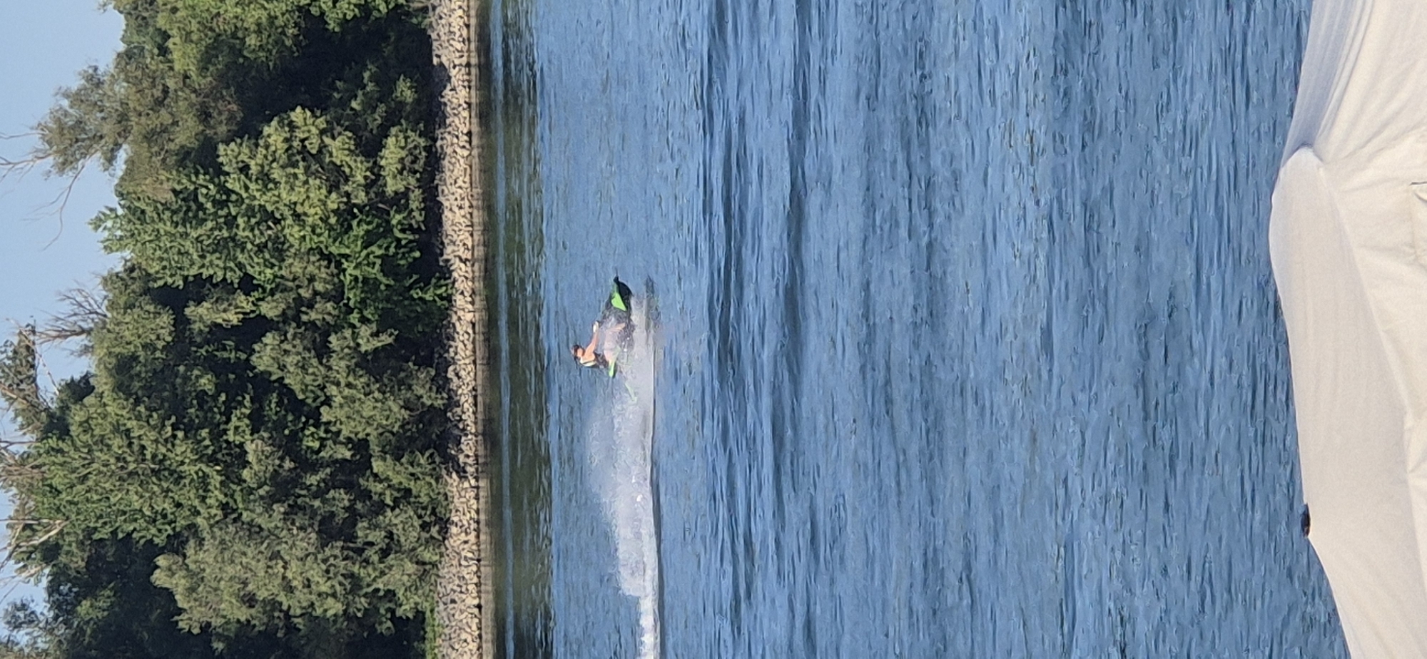 A person is riding a jet ski on a body of water. The jet ski is green and black, and the person is wearing a life jacket. The water is blue and choppy, with white spray coming off the back of the jet ski. In the background, there is a line of trees on a rocky shore. The scene appears to be a sunny day, possibly in the city of Mainz, Germany, given the vegetation and body of water commonly found along the Rhine River. In the foreground, the edge of a white object, possibly a boat cover or umbrella, is visible.