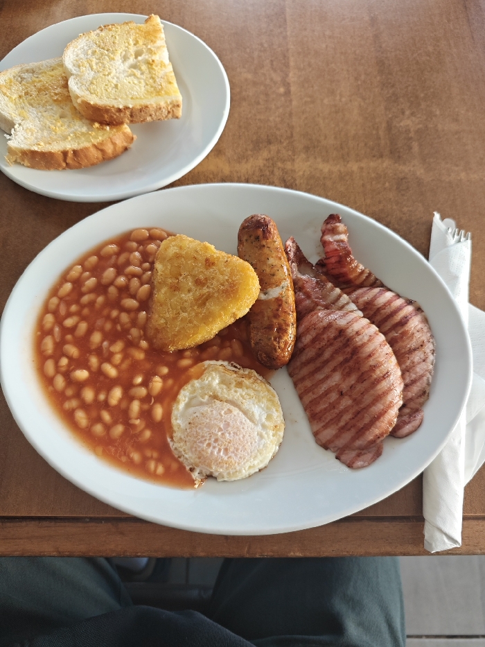 An overhead view captures a dining scene at a table in a dining establishment in London, United Kingdom. The primary subject is a meal arranged on two white ceramic plates on a light brown wooden tabletop. The larger, oval-shaped plate in the foreground holds a "Full English Breakfast" consisting of a substantial portion of baked beans in tomato sauce, a single heart-shaped golden-brown hash brown partially submerged in the beans, one browned pork sausage, three grilled rashers of back bacon with prominent grill marks, and a fried egg with a firm white yolk and slightly crisp edges, also partially in the beans. To the upper left, a smaller circular white plate contains two slices of lightly toasted bread, buttered or margarined. A rolled white paper napkin and the tines of a metallic fork are visible to the right of the main plate. The lower edge of the frame shows the dark trousers of a seated individual. The lighting is bright and even, indicating an indoor setting.