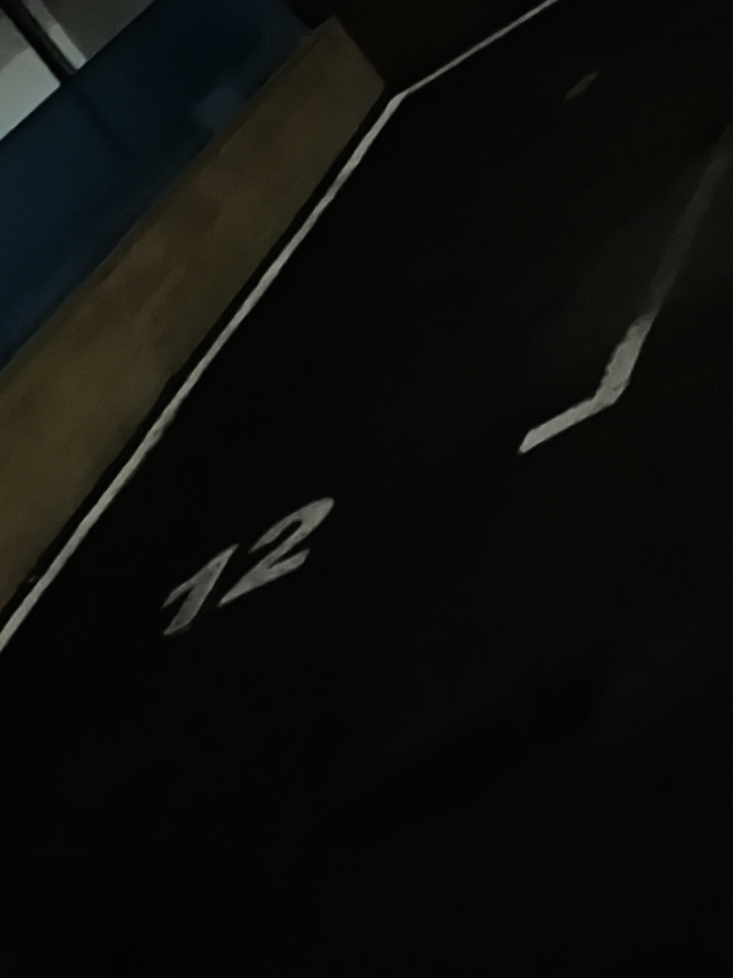 The image shows a parking spot in what appears to be an indoor garage, labelled "12". The spot is marked with white lines and the number is painted in white. The light is dim, creating shadows. A partial view of a wall or structure is visible to the left, with dark blue and tan sections. No people are visible. It is impossible to verify the location based on the image itself.