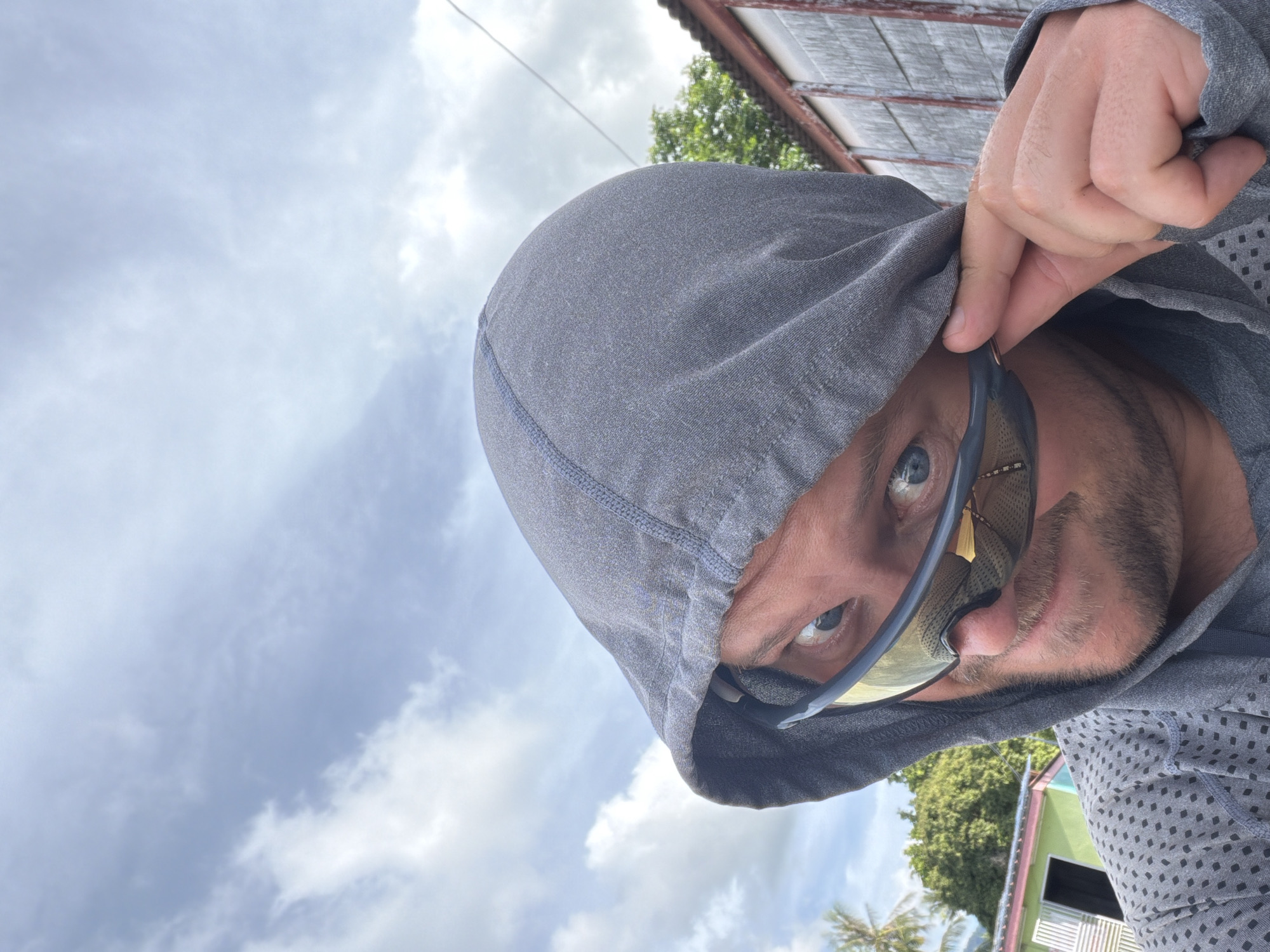 A man wearing a grey hooded jacket and sunglasses is taking a selfie outdoors. The man is looking directly at the camera with a playful expression. He is adjusting his sunglasses with his right hand. In the background, there are some buildings and trees, suggesting an outdoor location in a tropical or sub-tropical climate. The sky is partly cloudy. The style of the buildings in the background and the presence of palm trees indicate a possible location in Southeast Asia, such as Baan Mae Nam, Thailand.