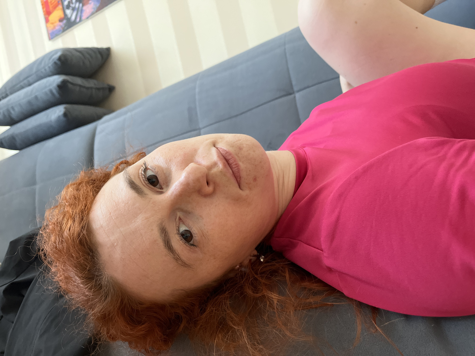 A woman with auburn hair is lying on a gray sofa. She is wearing a pink top. There are gray pillows stacked on the left side of the sofa. A picture hangs on the wall behind the sofa. The location is Chaikovsky, Russia.