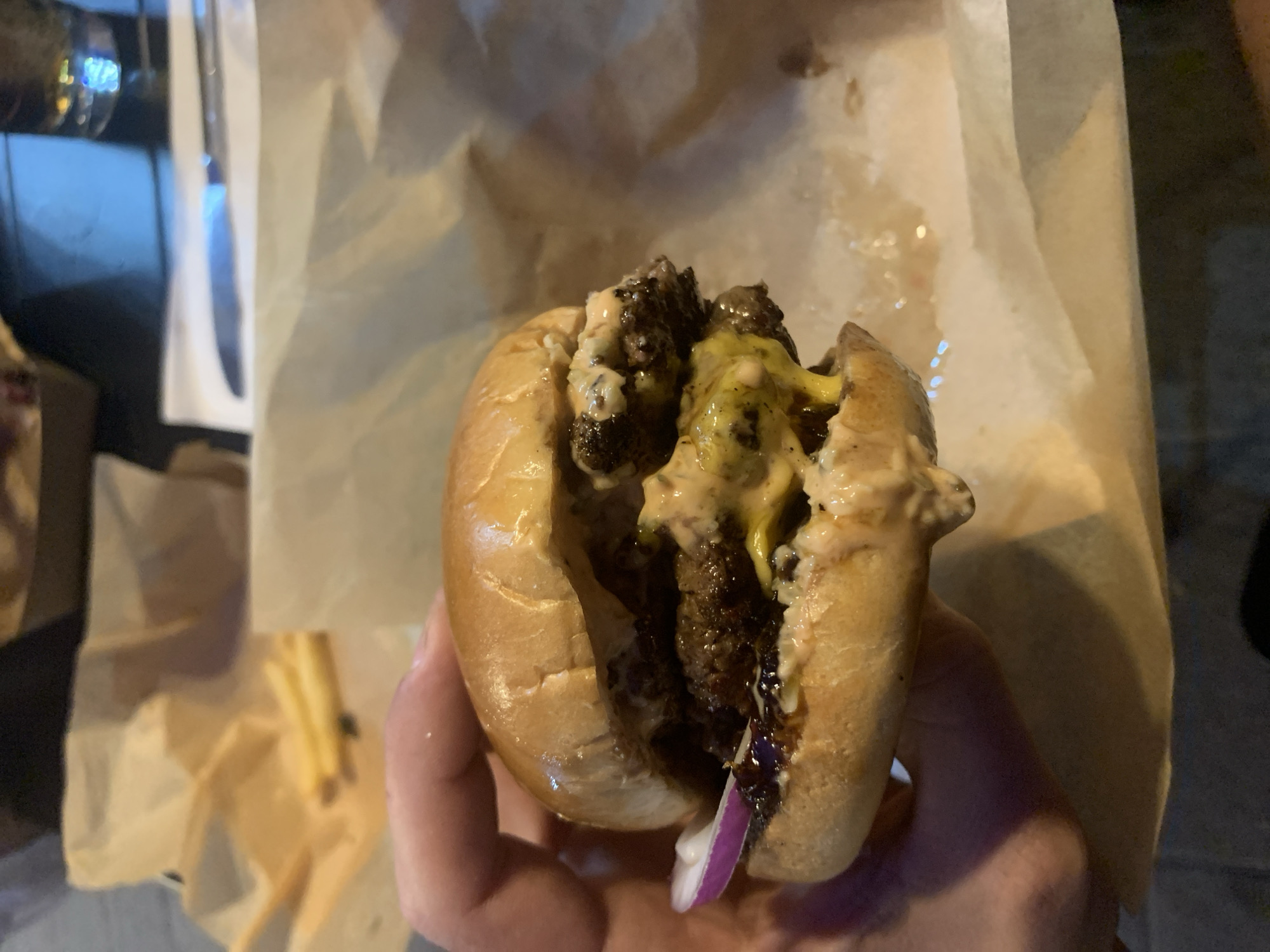 A close-up shot of a double-patty burger being held in someone's hand, presented on a piece of parchment paper with a few scattered fries visible in the background. The burger features two beef patties, melted cheese, sauce, and a slice of red onion. The bun appears to be a soft, glazed type. The lighting suggests an indoor setting, possibly a restaurant or food stall. While the exact location within Tel Aviv, Israel cannot be determined from the image alone, the burger's presentation suggests a casual dining experience in a modern urban environment.