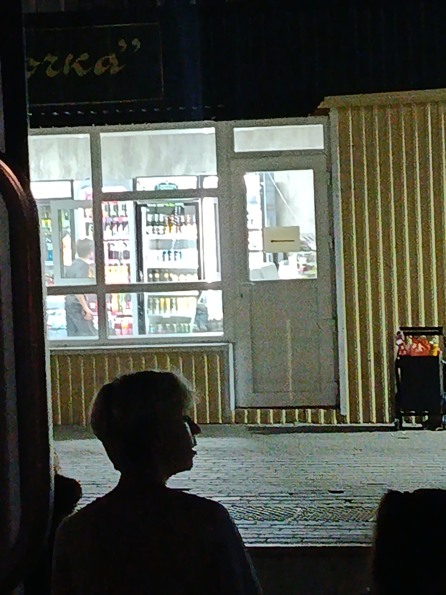The image depicts a storefront in Rtishchevo, Russia, likely a small shop or kiosk, at night. Above the storefront, a dark sign with stylized yellow lettering is visible, with the visible portion reading "чка'". The storefront features large glass windows revealing the interior, which is brightly lit. Inside, shelves are stocked with bottles, suggesting it might be a beverage or convenience store. Two individuals are partially visible through the window: one standing and looking into the store, and another inside the store, possibly an employee, interacting with items on the shelves. The storefront is attached to a building with a corrugated metal exterior in a light brown or beige color. To the right of the storefront, a dark trolley cart with some items, possibly merchandise, is present. In the foreground, the silhouettes of at least two people are visible, suggesting they are standing outside the storefront, looking in or passing by. The ground appears to be paved.
