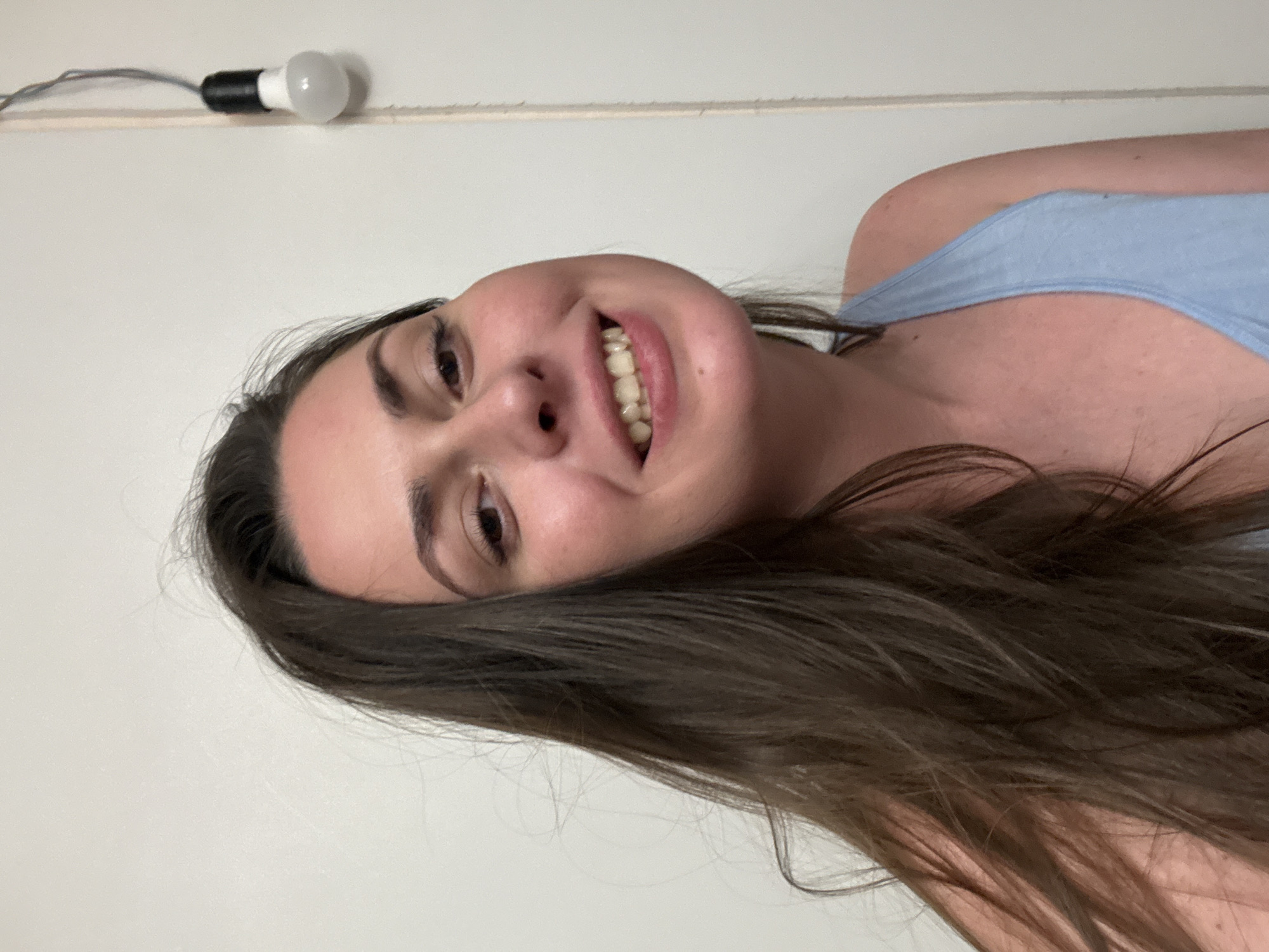 A young woman with long, dark brown hair smiles widely at the camera. She is wearing a light blue tank top. Her skin is fair and appears slightly flushed. Her eyes are dark brown and her eyebrows are well-defined. The background is a plain white wall. To the right of the woman, a bare lightbulb hangs from a cord, attached to the wall. The lighting is direct and somewhat harsh, creating shadows on the woman's face. The image is a close-up, focusing on the woman's head and shoulders.