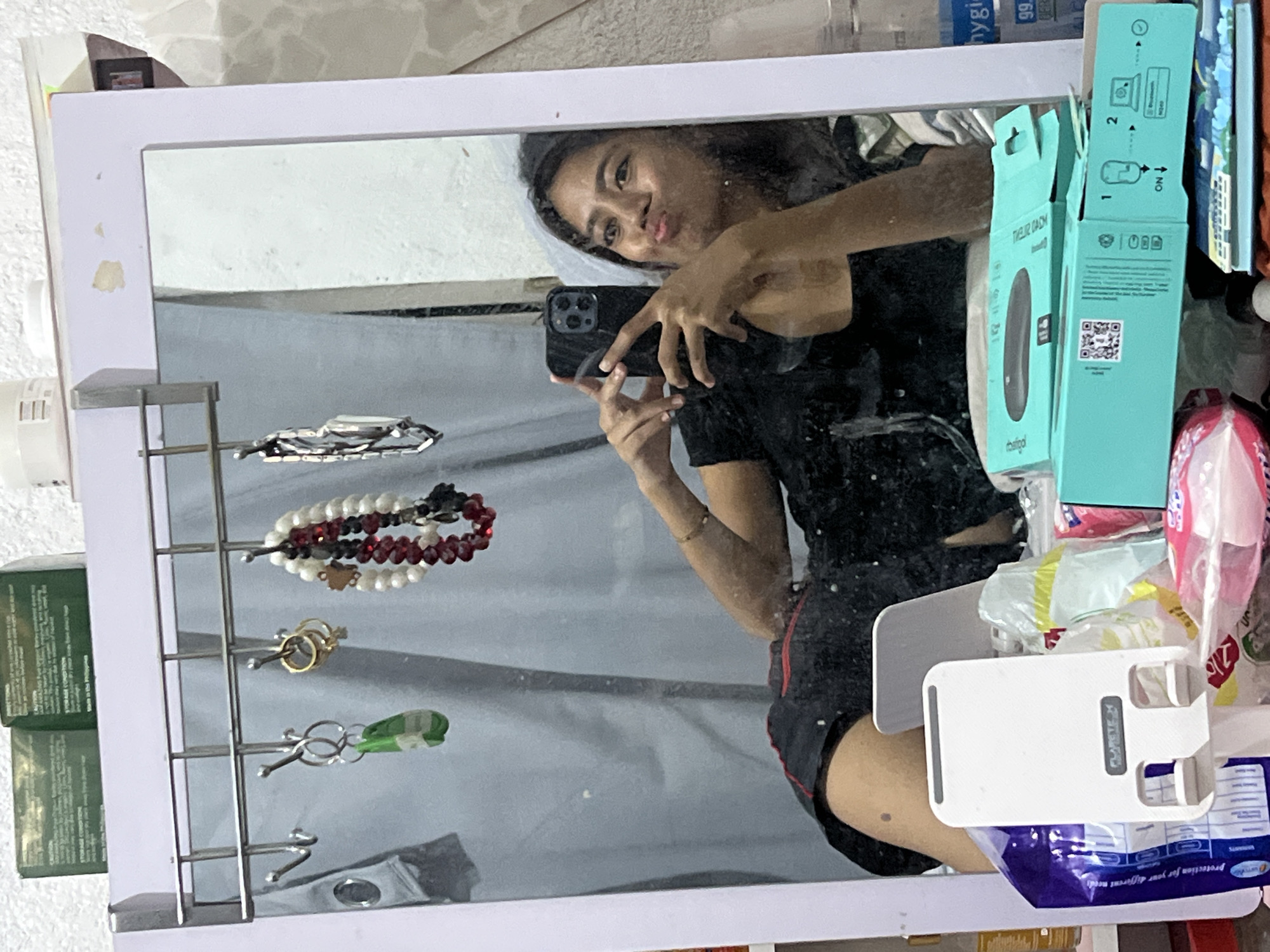The image is a mirror selfie taken in what appears to be a bedroom in Davao City, Philippines. A young woman with dark hair, wearing a black t-shirt and black shorts with red trim, is lying down and taking the picture with her smartphone. She is making a kissing face.
In the foreground, to the left, is a white phone stand with the brand name "FLARETECH" visible. Behind the stand are various items, including a purple package, a plastic bag with yellow and white accents, and a teal box for a computer mouse.
The mirror reflects a hanging rack above, on which several items are displayed: a green object on a keyring, a set of rings, and two beaded necklaces, one white and the other red and black. Behind the rack, a gray curtain is visible. The background of the room shows a white wall with some texture and possibly some items on a shelf above the mirror.