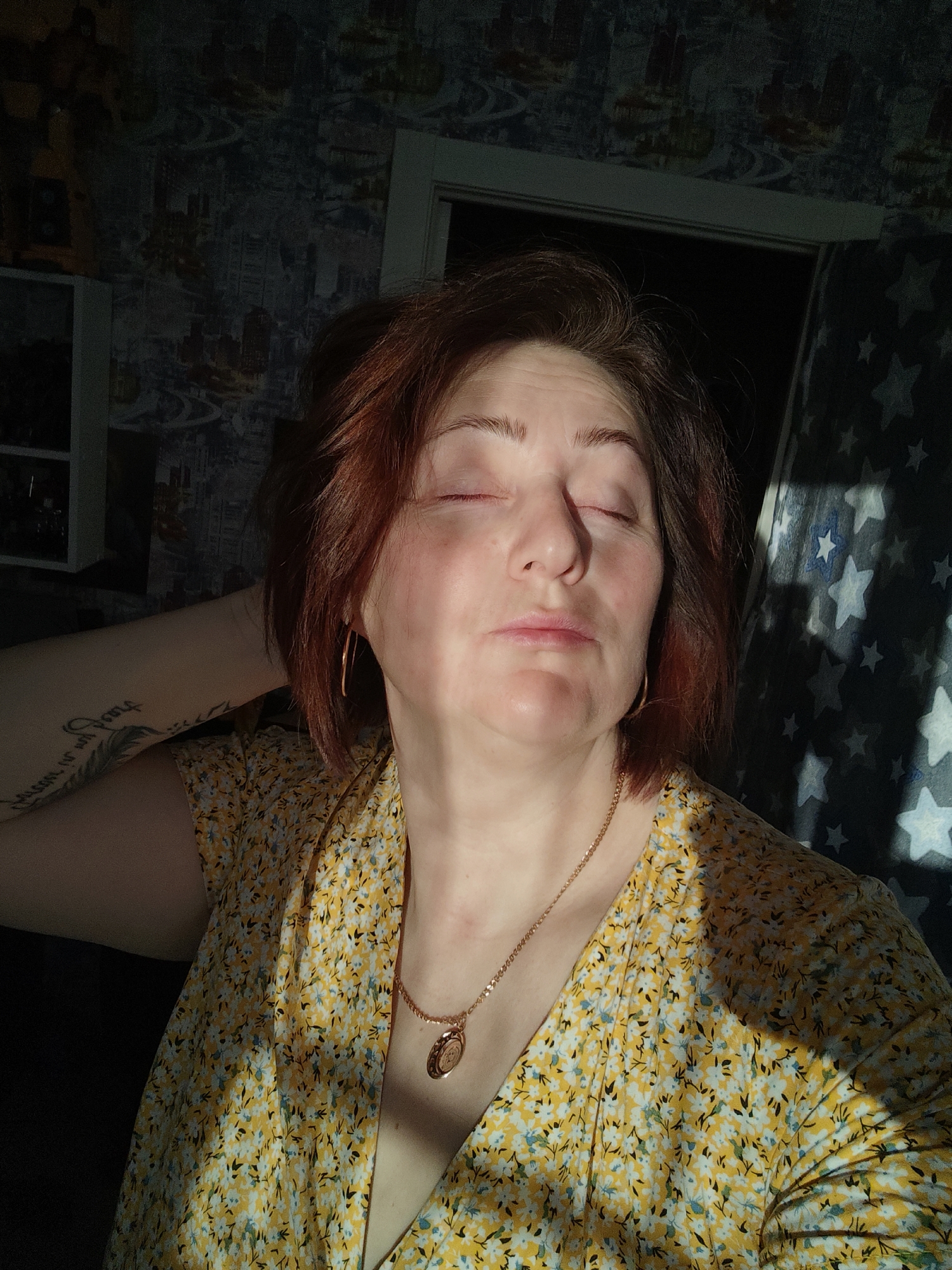 The image depicts a person with reddish-brown hair and closed eyes, possibly a woman, wearing a yellow floral print dress and gold jewelry including hoop earrings and a necklace with a pendant. Part of an arm with a tattoo is visible behind her head. The background consists of patterned wallpaper and a partially obscured window covered with a star-patterned curtain. A shelf unit can be seen to the left of the individual. The strong, direct sunlight suggests it is daytime. No specific details confirming the exact location in Markovskii, Russia are visible.