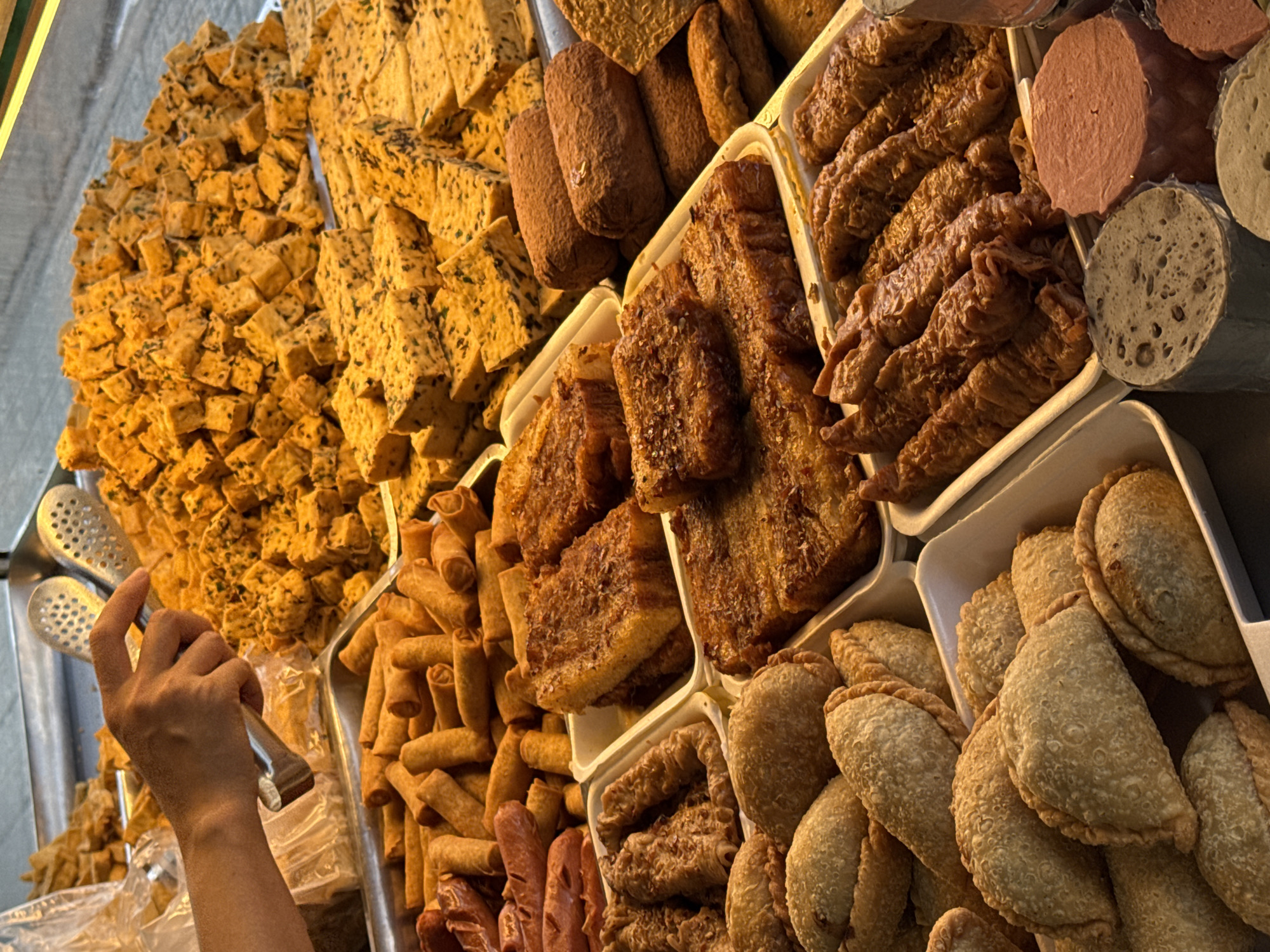 This vertical shot captures a vibrant street food stall, likely at a market in Da Lat, Vietnam, displaying a wide array of prepared savory snacks. The scene is brightly lit by daylight, suggesting a daytime market setting.
The stall features numerous food items arranged in white plastic trays and metal containers. Prominently in the upper left, a large quantity of small, golden-brown, cubed items, possibly fried tofu or a savory cake (like "bánh đúc mặn"), are piled high. These pieces are speckled with dark green herbs.
Towards the center and right, other fried delicacies are visible. There are long, dark brown cylindrical items and flatter, textured rectangular pieces, possibly various types of fried dough or cakes. Numerous golden-brown, rolled items, resembling mini spring rolls or "chả giò," are stacked in a tray. Further right, a collection of half-moon shaped, deep-fried pastries with crimped edges, similar to empanadas or "bánh gối," are neatly arranged.
Also visible are several processed items: reddish-brown sausage-like pieces and round, pale discs, some whole and others cut to reveal a porous, textured interior, characteristic of certain Vietnamese pork sausages or vegetarian alternatives.
In the mid-left of the frame, a human hand is holding a pair of metal tongs, poised as if to serve or select food. The action suggests a transaction at the stall, with customers choosing their desired items. The overall impression is one of abundance and variety, typical of Vietnamese street food culture. There is no visible text.