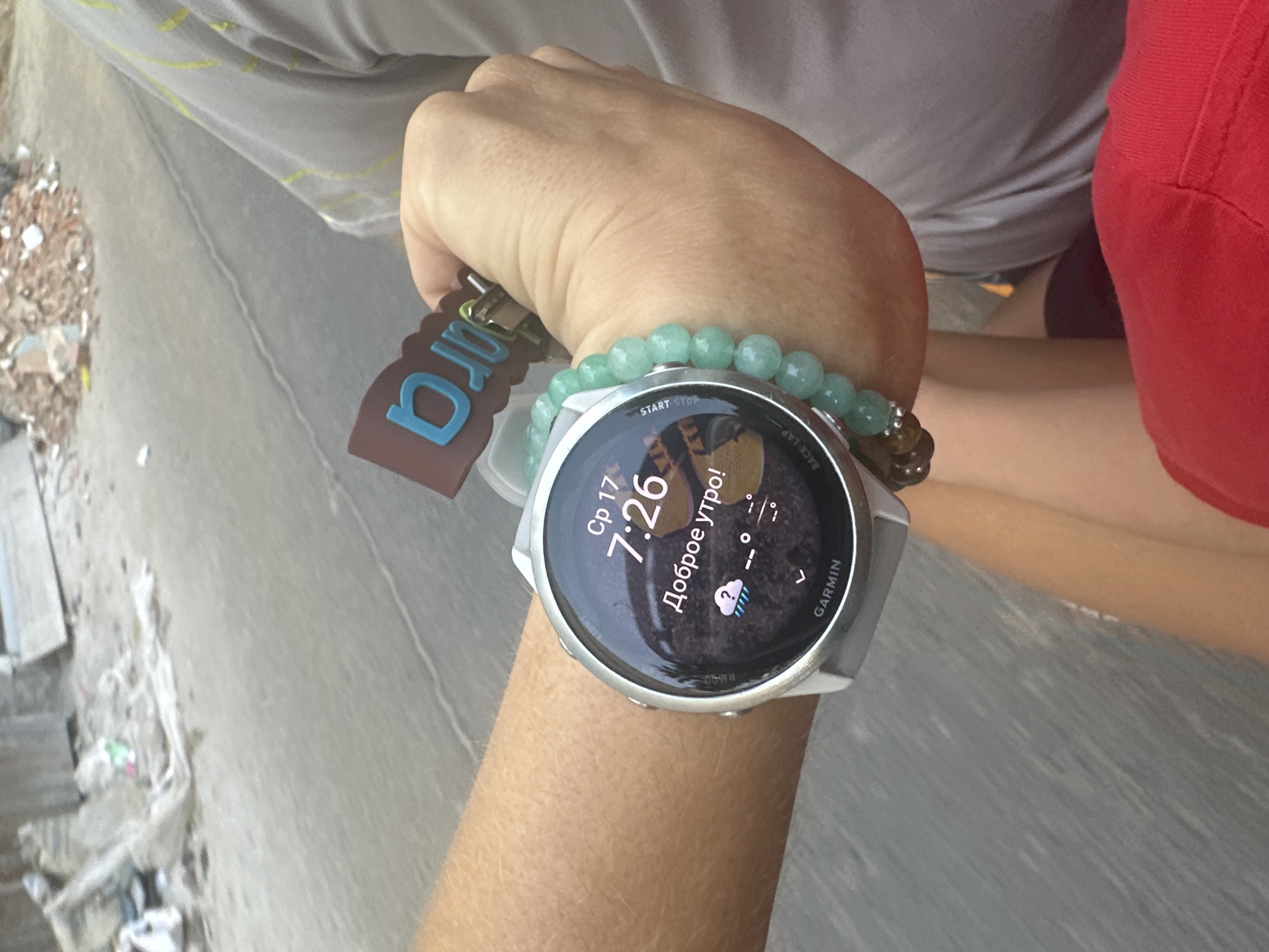 The image shows a close-up of a person's left wrist wearing a grey Garmin smartwatch and a bracelet made of light green beads. The smartwatch screen displays the time "7:26", the date "Cp 17", and the greeting "Доброе утро!" (Good morning!) in Russian. A small weather icon depicts rain and clouds. Attached to the watch strap is a brown tag with the blue letters "DJ". The background is blurry and appears to be an outdoor setting with a grey asphalt surface and some debris, possibly construction waste, in the upper portion of the frame. The environment suggests an urban or suburban location.