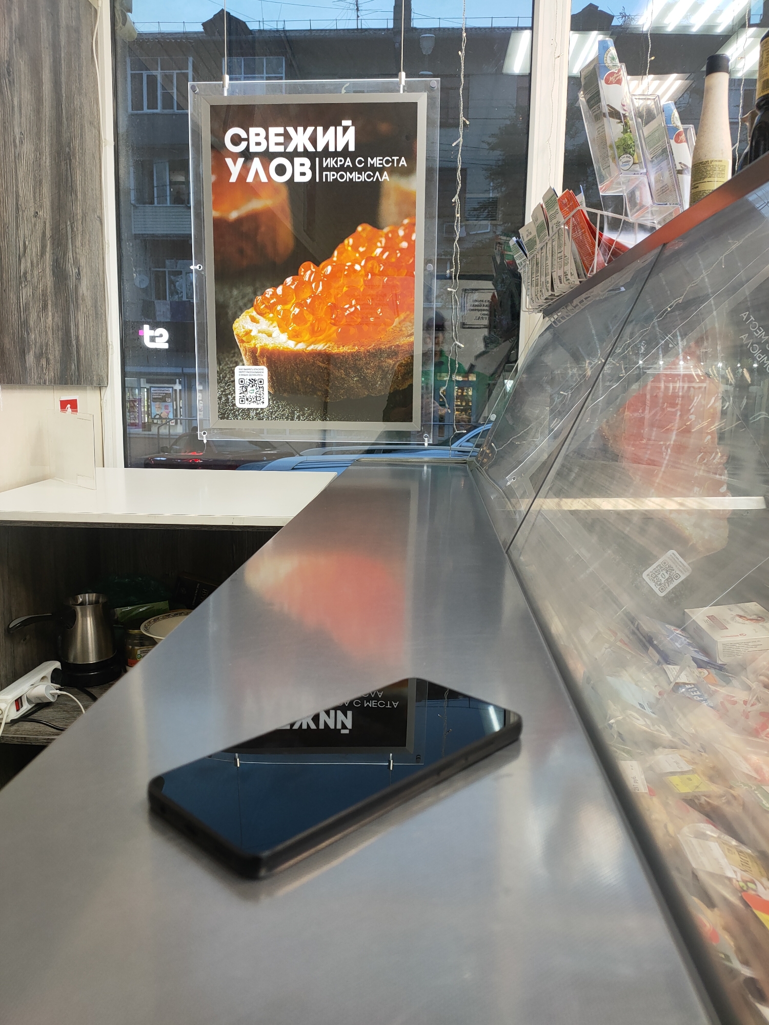 The image presents an interior view of a retail space, likely a food shop specializing in seafood, set in an urban environment. A metallic counter surface, reflecting overhead lights and parts of the room, occupies the foreground. A black smartphone lies face-down on this counter, its screen reflecting reversed text.
Behind the counter, a shelf holds an electric kettle, a white power strip plugged in, a plate, and some canned goods. A large window forms the back wall, through which an apartment building and other distant structures are visible, suggesting a daytime scene in a city. A "t2" logo, likely belonging to the Russian mobile operator Tele2, is visible through the window.
A prominent, clear acrylic sign is suspended in front of the window, advertising "СВЕЖИЙ УЛОВ" (Fresh Catch) and "ИКРА С МЕСТА ПРОМЫСЛА" (Caviar from the Fishing Grounds). The sign features an appealing image of bright orange caviar on a piece of bread and includes a QR code at its base.
To the right, a long, clear display case, presumably for chilled goods, runs alongside the counter. Inside the case, various packaged products are visible, with one label clearly showing "297 руб." (297 rubles). Decorative string lights are draped above this display. The reflective surfaces throughout the image, including the phone and display case, show fragmented, reversed text from the surroundings. The scene is static, with no people visible or specific activity taking place. The location is Sochi, Russia.
