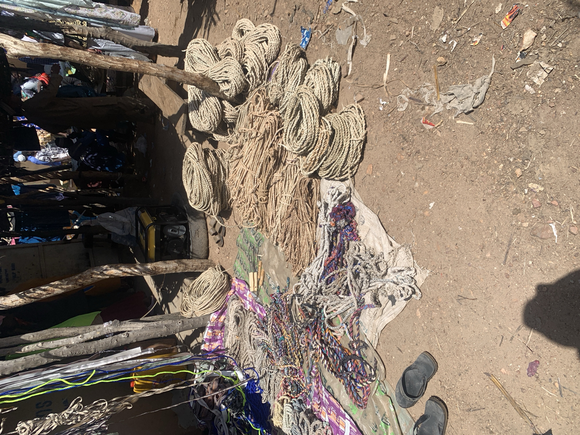 This media file captures an outdoor scene in a marketplace or informal trading area, likely in Nigeria, given the project context. The primary subjects are a large collection of ropes and cords of various thicknesses, colors, and materials, all piled on the ground, some on what appears to be a tarp or fabric. The ropes range from thick, natural fiber ropes bundled together, to thinner, brightly colored synthetic cords, including shades of blue, green, and purple. Some of these are interwoven, suggesting they might be for sale as decorative items or for practical use.
In the background, a rough wooden structure with additional ropes and what looks like fabric or clothing draped over it is visible. A yellow generator is also present in this area. Scattered on the dirt ground are debris, including pieces of plastic and other small objects, indicative of an outdoor, possibly dusty environment. Two dark-colored flip-flops are placed on the ground near the bottom right of the frame, suggesting someone was recently present or is currently in the vicinity. The lighting suggests it is daytime, with clear shadows indicating the sun is at a moderate height. There is no visible text in the image. The overall impression is of a busy, possibly informal trading post where various goods, particularly ropes and cords, are being displayed and sold.