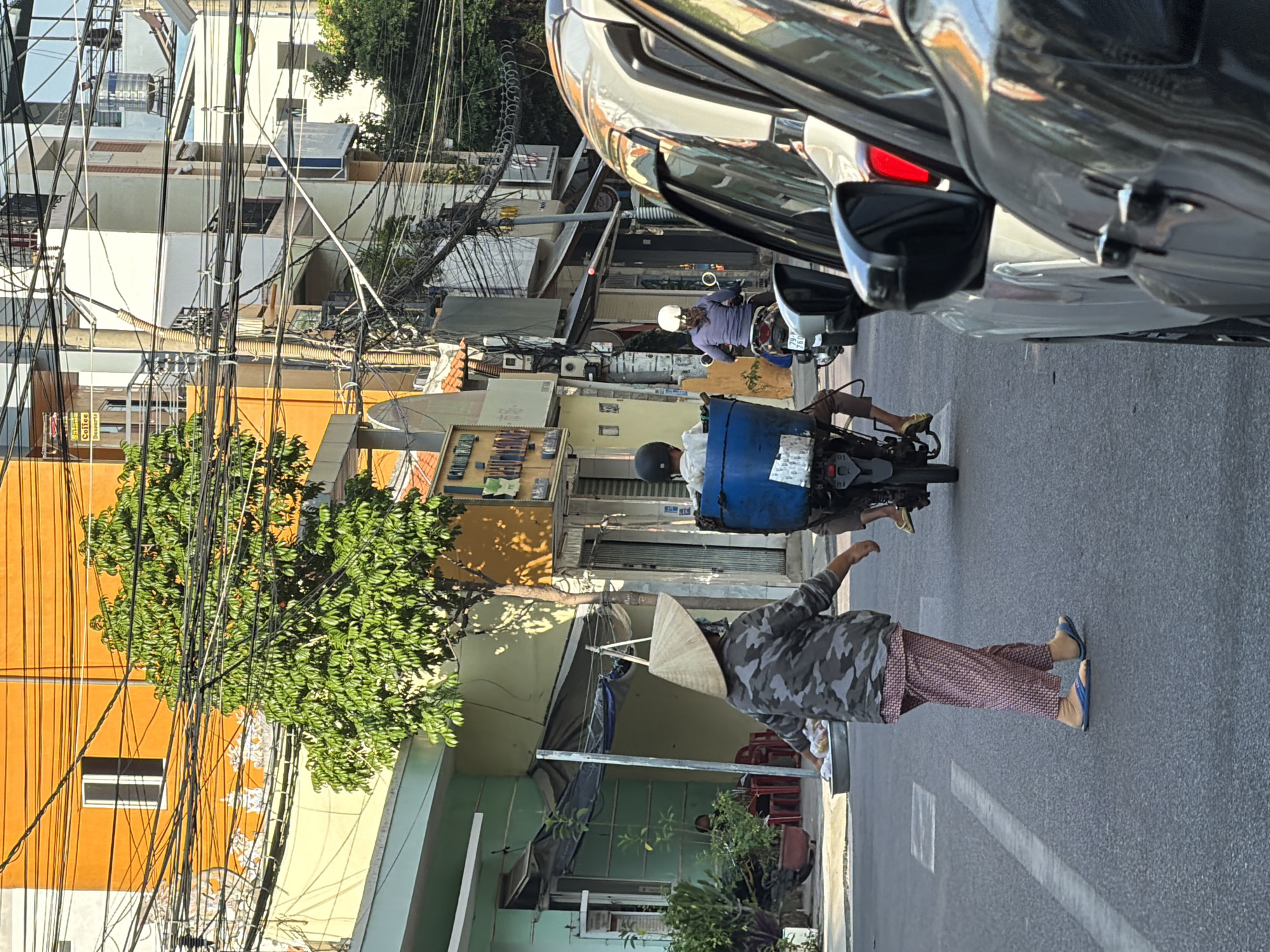 This is a street scene in Nha Trang, Vietnam, likely in the mid-morning based on the strong shadows. The foreground shows the side of a dark-colored car, partially obscuring the view. To the left of the car, another vehicle, possibly white, is visible, with a red brake light illuminated.
In the mid-ground, a person wearing a conical hat and a camouflage-patterned jacket is walking from right to left across the street. They are also wearing patterned trousers and blue flip-flops, and are carrying a round metal tray containing what appear to be food items.
Behind this person, two motorcycles are present. The first motorcycle, closer to the foreground, has a rider wearing a helmet and carrying a large blue plastic container on the back. The rider's legs are visible, wearing light brown trousers and yellow footwear. Further back, a second motorcycle is seen with a rider wearing a white helmet and a purple jacket.
The background features buildings with vibrant yellow and orange facades, as well as green and white structures. Numerous electrical wires are strung haphazardly across the scene, a common sight in Vietnamese cities. A tree with lush green foliage stands near the center of the frame, casting shadows on the street. The street itself is paved with asphalt and has white lane markings. A few potted plants are visible near a building on the left. The overall impression is of a busy urban street with typical local activities.