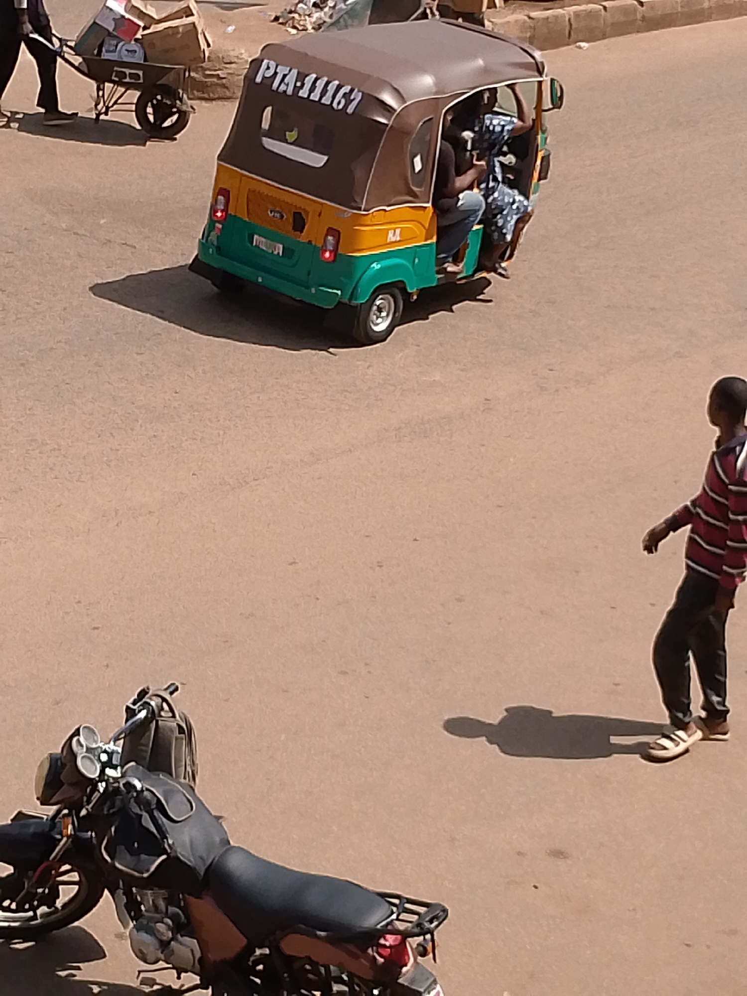 In an outdoor, sunlit urban environment, a three-wheeled vehicle, possibly a tuk-tuk or auto-rickshaw, with a brown canopy and a green and yellow body, is parked on an asphalt road. The vehicle's license plate reads "PTA-11167". Inside the vehicle, two individuals are visible: one appears to be a driver, and the other is a passenger wearing a patterned dress. To the left of the vehicle, a wheelbarrow laden with cardboard boxes and other items is being pushed by a person partially visible at the edge of the frame. In the middle ground, a young person wearing a striped long-sleeved shirt and dark pants walks away from the camera. In the foreground, the front of a parked motorcycle is visible. The overall scene depicts a street in an urban setting with various modes of transportation and pedestrian activity.