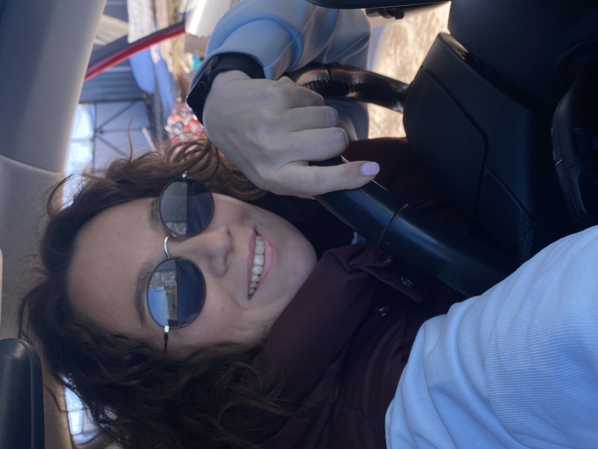 A woman with curly brown hair is wearing round, dark sunglasses and smiling. She is sitting in a car and holding the steering wheel. She is wearing a light blue, puffy jacket over a maroon button-down shirt. The image appears to be taken from inside the car, with a partial view of the exterior visible through the windows. The location is Ольховка, Russia.