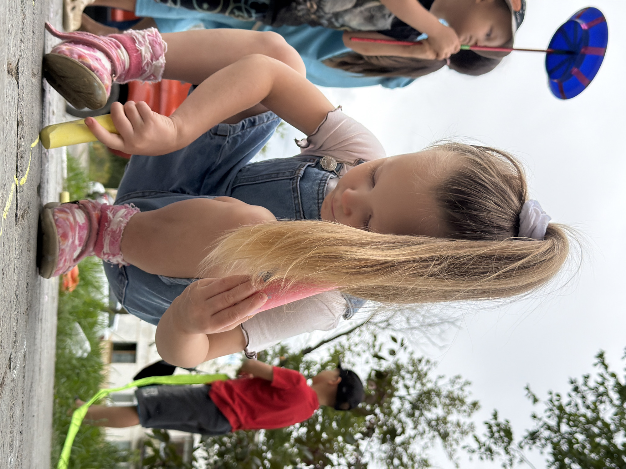 A low-angle, eye-level outdoor shot captures a young girl drawing with chalk on a paved surface. She is wearing denim overalls over a white t-shirt, and pink boots with white fur trim. Her blonde hair is pulled up in a high ponytail with a white scrunchie. She holds a yellow piece of chalk in her right hand, and a pink piece in her left. Her gaze is directed downwards at the chalk art she is creating. In the background, other children are visible, one holding a blue spinning toy, and another in a red shirt. Green grass and trees line the right side of the frame. The scene appears to be in an outdoor public space, potentially a park or a street, in Chaikovsky, Russia.
