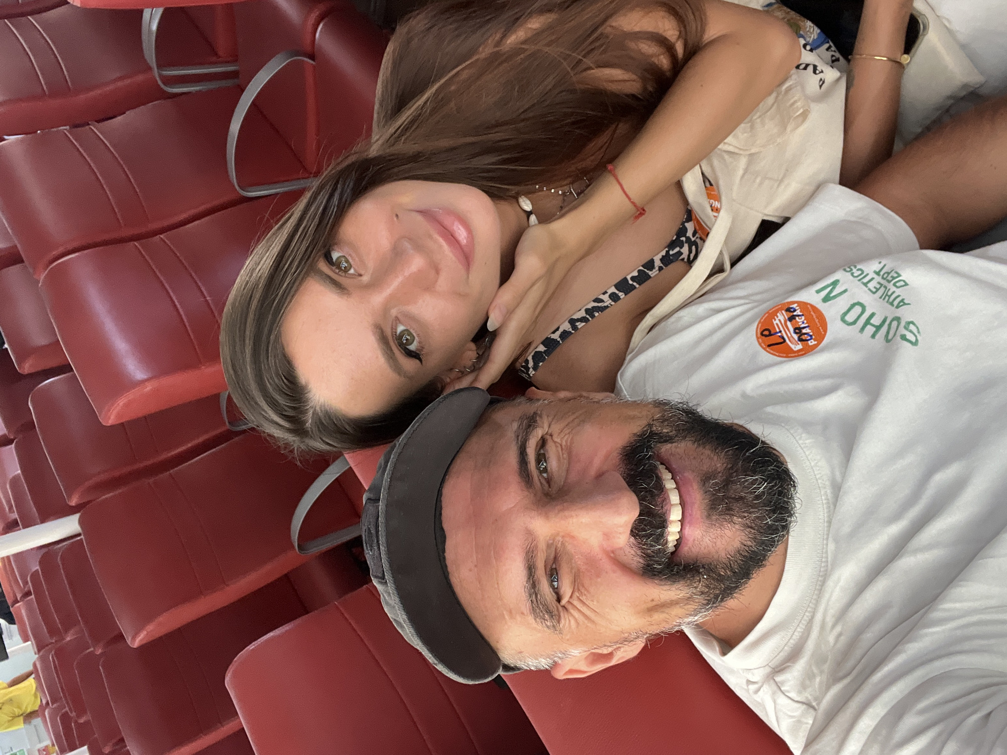 In Baan Bang Rak, Thailand, a man and a woman are taking a selfie in a ferry with rows of red seats. The man in the foreground wears a grey baseball cap and a white t-shirt with a circular logo and text that reads "SOHO N ATHLETICS DEPT.". He has a beard and is smiling at the camera. The woman, positioned behind and to the right of the man, has long brown hair and is looking at the camera with a gentle smile. She is wearing a leopard-print top and several necklaces, including one with beads and another with a pendant. A red string bracelet is visible on her wrist. A beige canvas bag is draped over her shoulder. The background consists of the red upholstered seats of the ferry.
