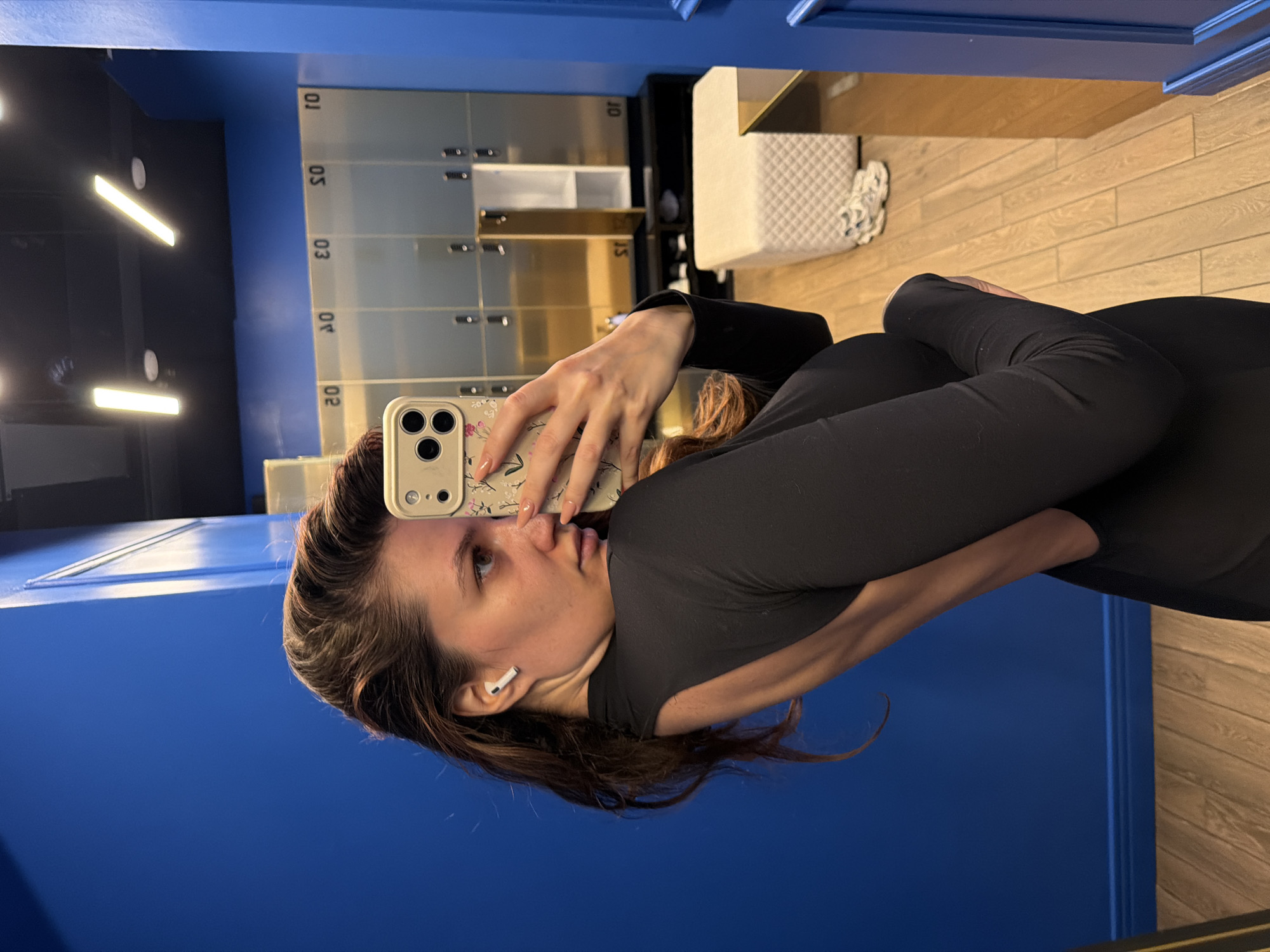 A woman is taking a selfie in a gym locker room. She is wearing a black long-sleeved shirt and black pants. She is holding her phone with a flowered case in her right hand and has an AirPod in her left ear. The locker room has blue walls and metallic lockers with numbers on them. A wooden bench can be seen in the background. The location of Belgrade, Serbia cannot be determined from the image.