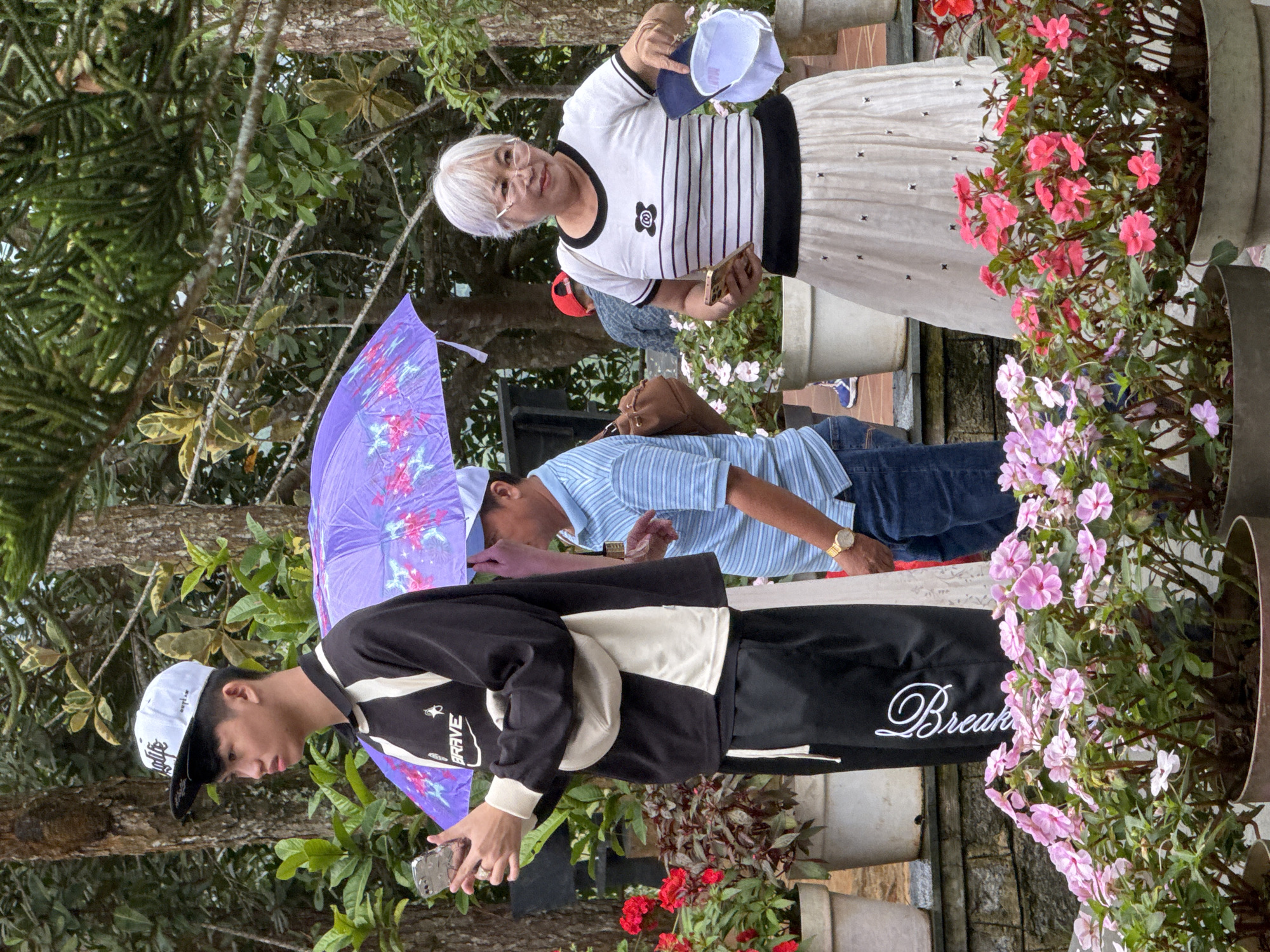 The image captures an outdoor scene with people and floral arrangements, likely in Da Lat, Vietnam. In the foreground, several potted plants with pink and purple flowers are visible. Behind them, a young person wearing a black and white long-sleeved shirt, black pants, and a white baseball cap is holding a smartphone. To their right, a man in a blue striped shirt and jeans is partially visible, holding a purple umbrella. Further to the right, a woman with white hair, glasses, and a black and white striped top paired with a long white skirt is also holding a smartphone and a blue baseball cap. The background features trees and greenery, suggesting a park or garden setting. The overall atmosphere appears to be casual, with people enjoying the outdoors.