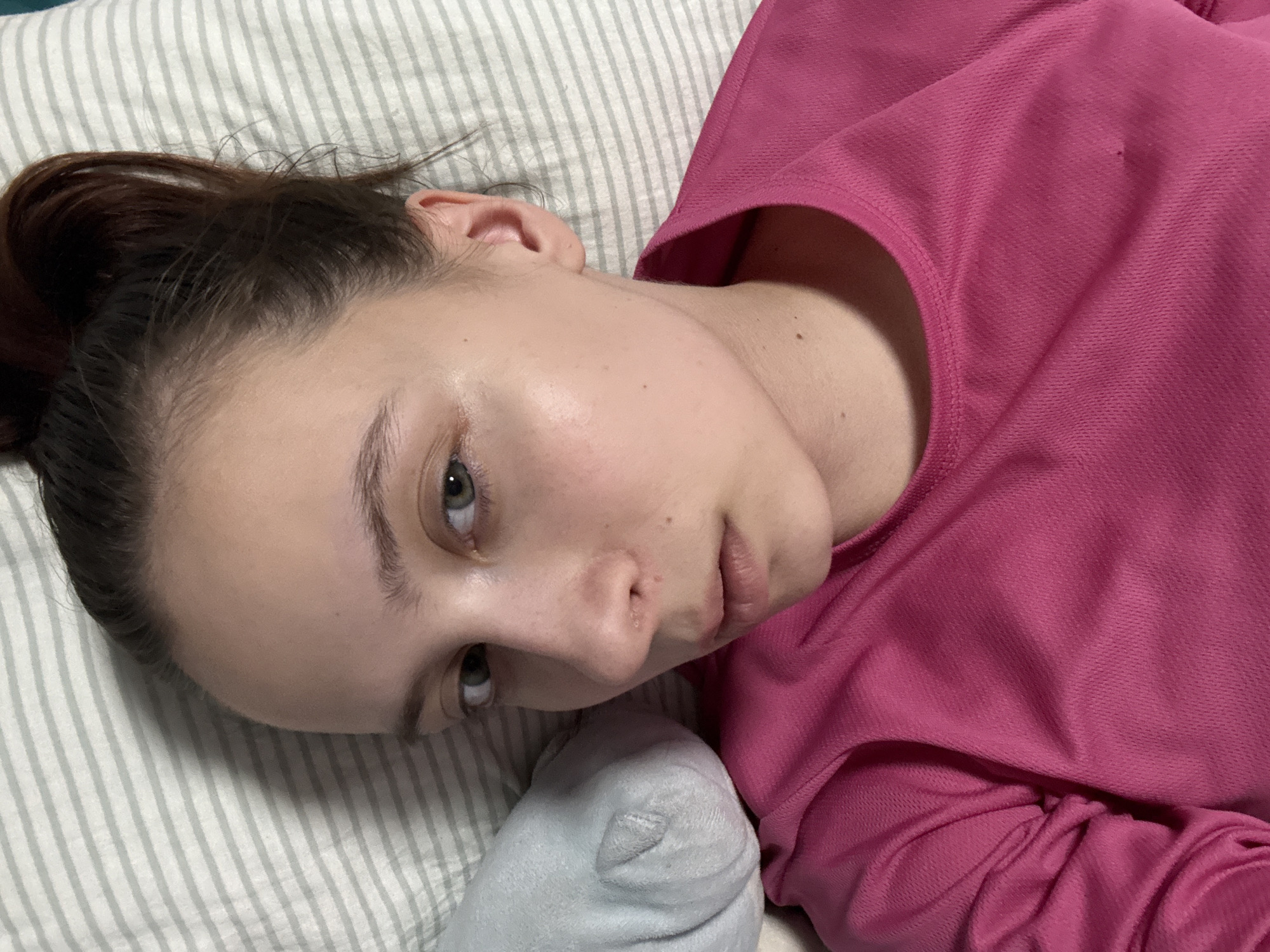 A person lies on a striped pillow. They wear a pink long-sleeved shirt. Their hair is pulled back. A gray sock rests near their face. The location is unknown, there is no evidence of Прикамский, Russia.