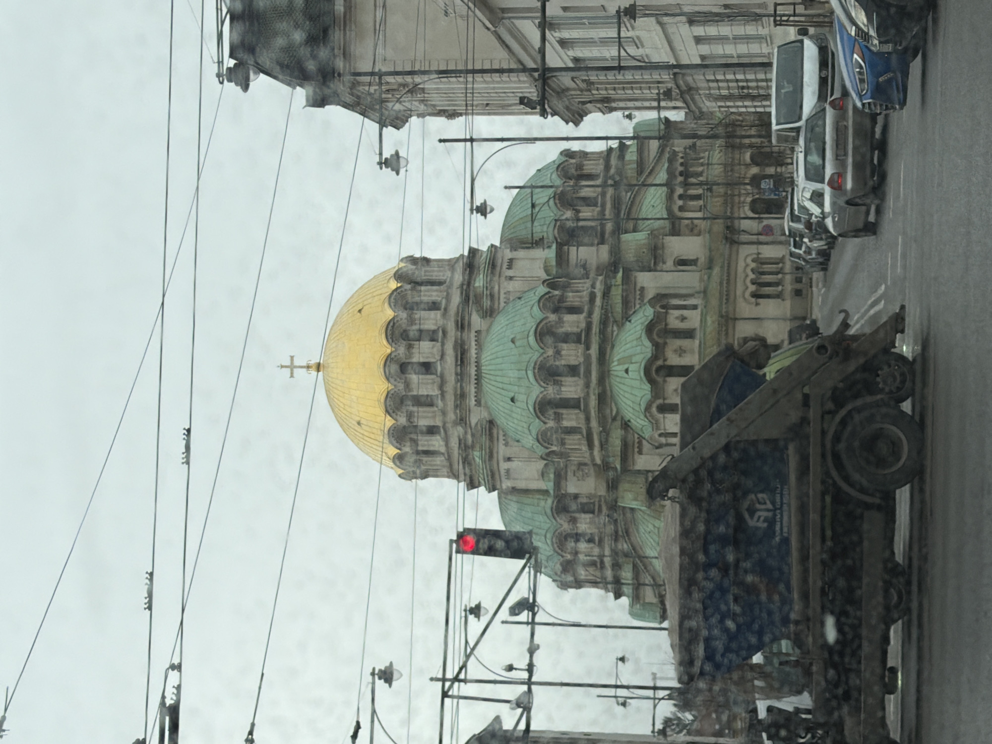 The image shows the Alexander Nevsky Cathedral in Sofia, Bulgaria, viewed from a street-level perspective on a cloudy day. The cathedral, with its prominent gold dome and green domes, is partially obscured by utility poles and wires. In the foreground, a truck and several parked cars are visible along the street. A red traffic light is illuminated. Buildings line the street on either side. The overall environment suggests an urban setting within the city.