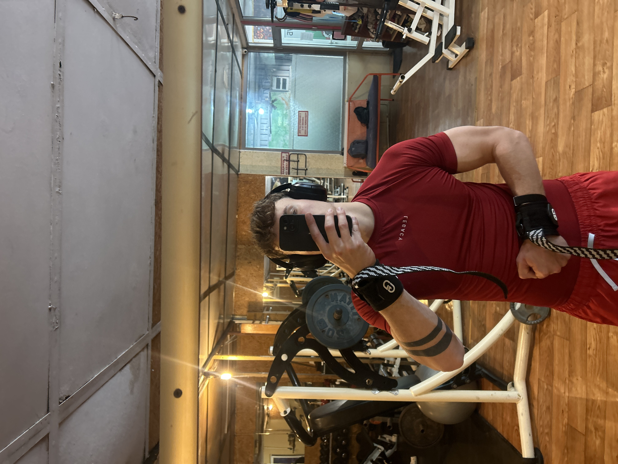 A man is in a gym, taking a selfie in a mirror. He is wearing a red t-shirt and red shorts. He has headphones on his head and wrist wraps on both wrists. He is holding a phone in his left hand. He is standing near a weight machine. Gym equipment is visible in the background. Through a window, a building can be seen. This image was taken in Elhovo, Bulgaria.