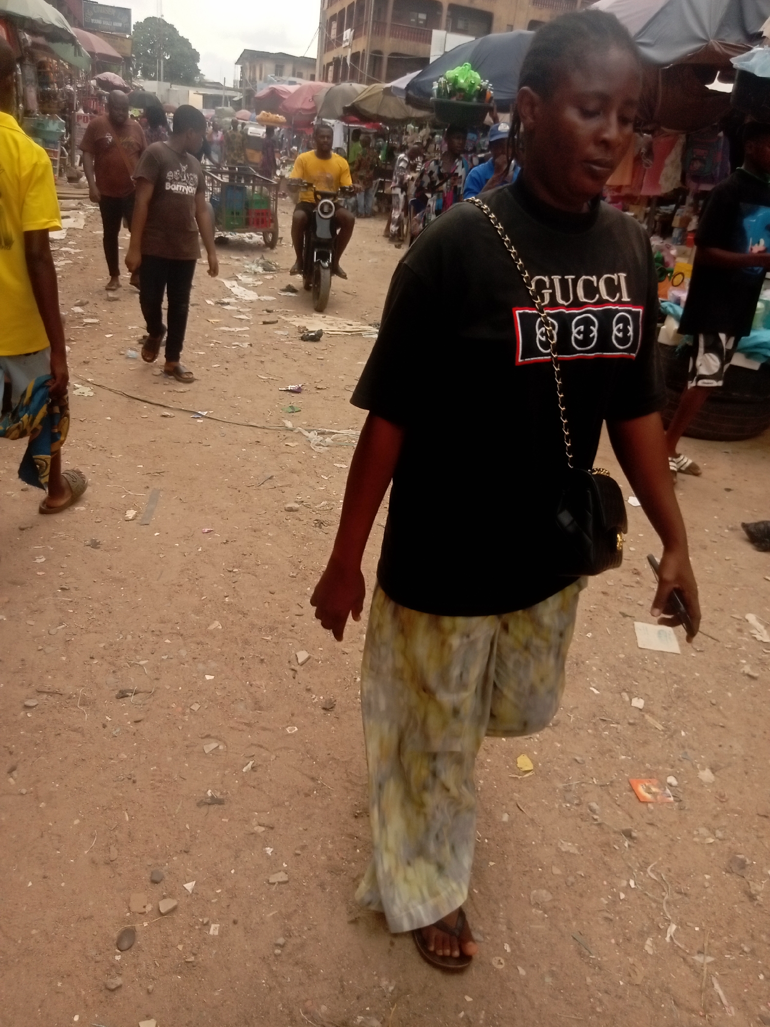 The image shows a bustling scene in Fegge, Nigeria, with a dirt road serving as a primary thoroughfare. In the foreground, a woman wearing a black Gucci t-shirt, patterned pants, and sandals is walking forward, holding a phone in her right hand and a small black purse hanging from a gold chain on her left shoulder. Behind her, several other people are visible, including one wearing a brown shirt and dark pants and another in a yellow shirt. A man on a scooter is navigating through the crowd. Vendors and their stalls are lining the sides of the road, with umbrellas providing shade. Litter, including paper and debris, scatters the ground. Buildings are visible in the background.