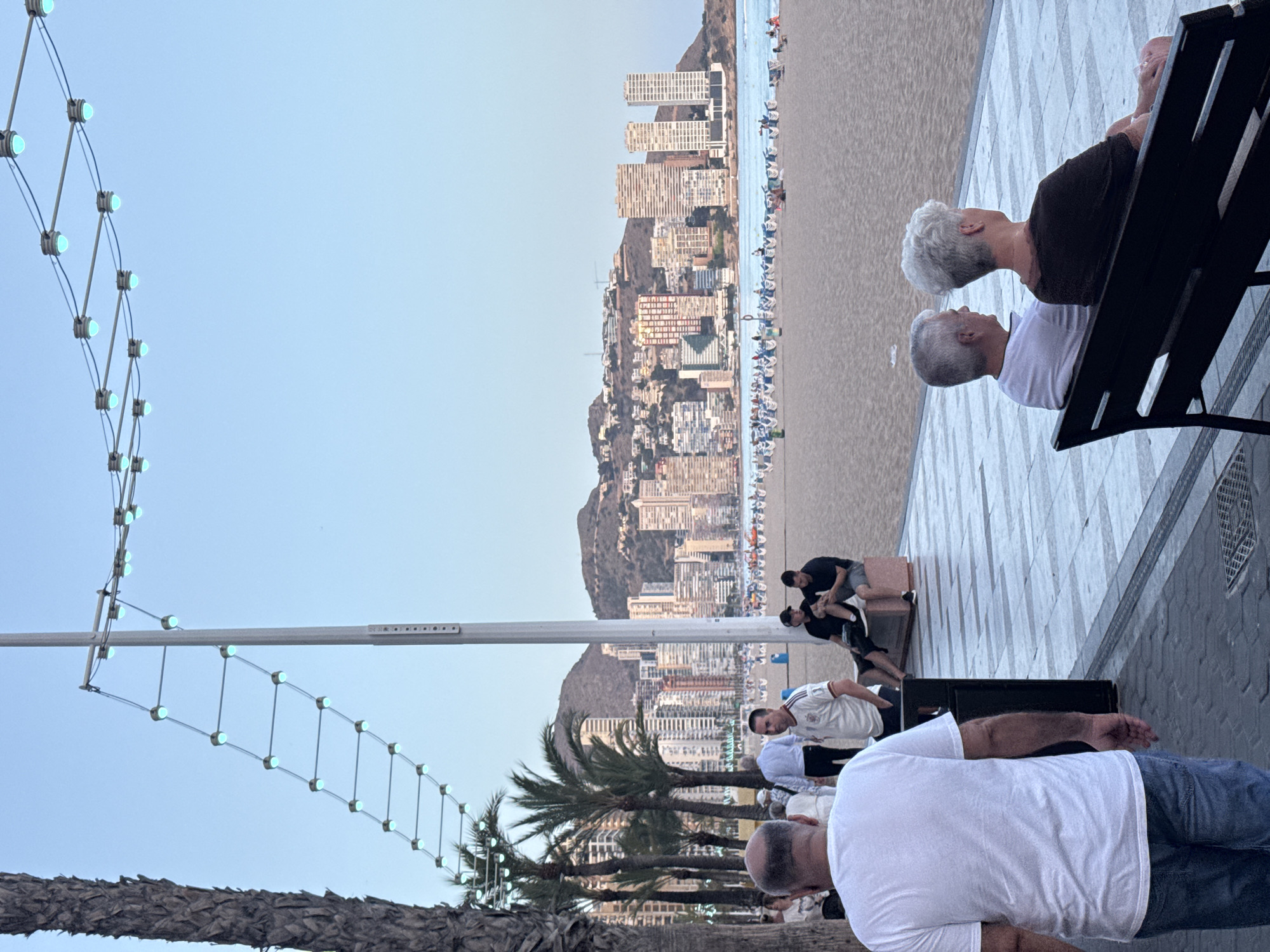 The image depicts a waterfront promenade in Benidorm, Spain, with the Mediterranean Sea and a sandy beach visible in the background. A row of palm trees separates the promenade from the beach, and beyond that, the cityscape of Benidorm rises, characterized by numerous high-rise buildings and a mountainous backdrop. String lights with blue bulbs are suspended above the promenade, contributing to the ambient lighting.
On the promenade, several individuals are present. In the foreground, a man with a shaved head wearing a white t-shirt and blue jeans is walking away from the camera. To his right, two men are seated on a bench, looking towards the sea. Further back, a man in a white and black sports jersey is standing. On the beach, lounge chairs are set up, and a few people are visible, some sitting and others walking. The sky is clear and light blue, suggesting it is late afternoon or early evening. The overall atmosphere is relaxed and suggests a popular tourist destination.