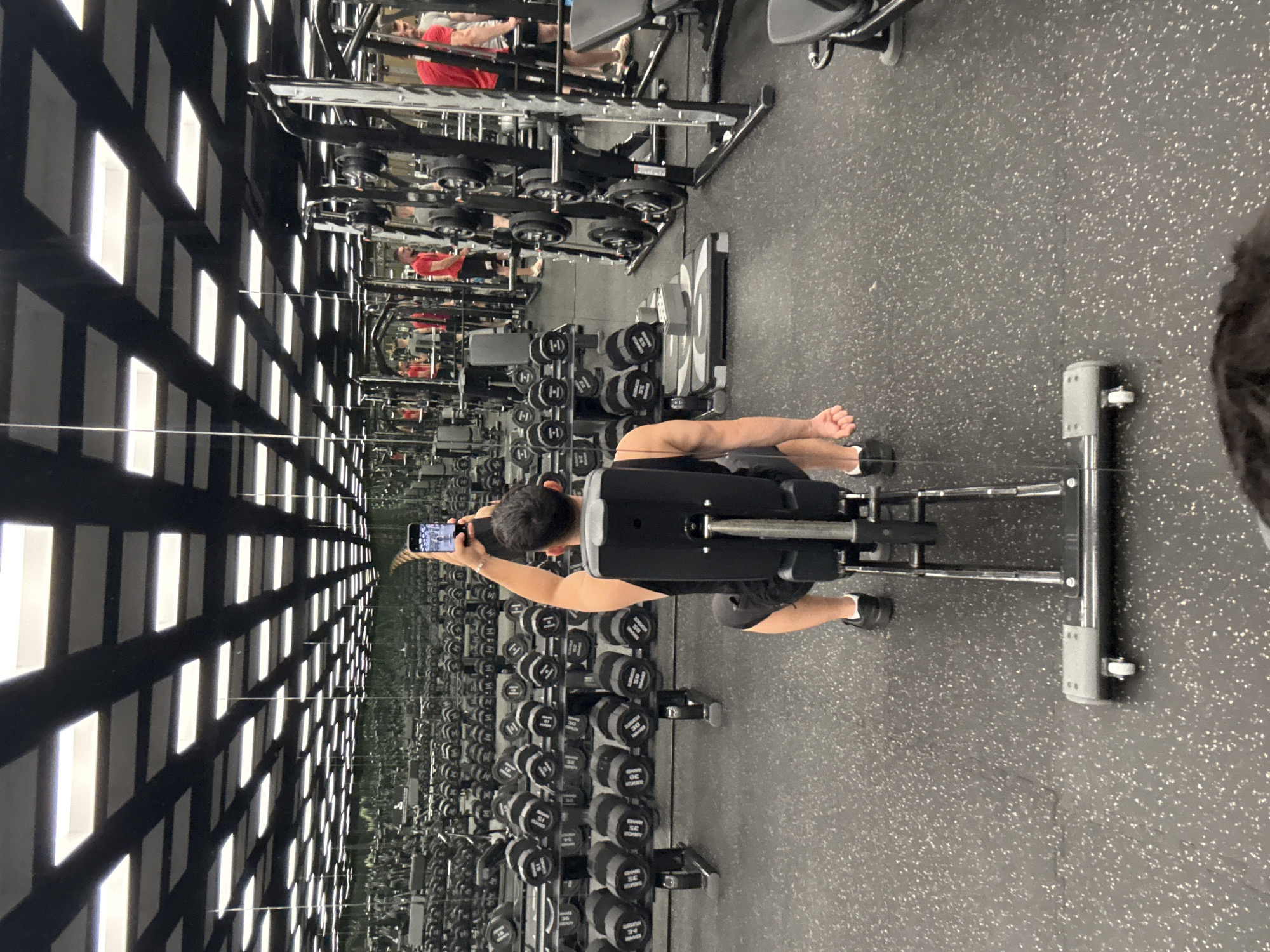 The image depicts the interior of a gym. In the foreground, a man is seated on a weight bench, holding a smartphone up to take a selfie. He is wearing a black tank top and black shorts. The gym is equipped with numerous weight racks filled with dumbbells, as well as various weight machines and exercise equipment. The ceiling features a grid of fluorescent light panels. Mirrors line the walls, reflecting the equipment and the man. In the background, other individuals can be seen exercising. The floor is covered with black rubberized material. The scene suggests a typical workout environment in a well-equipped gym.