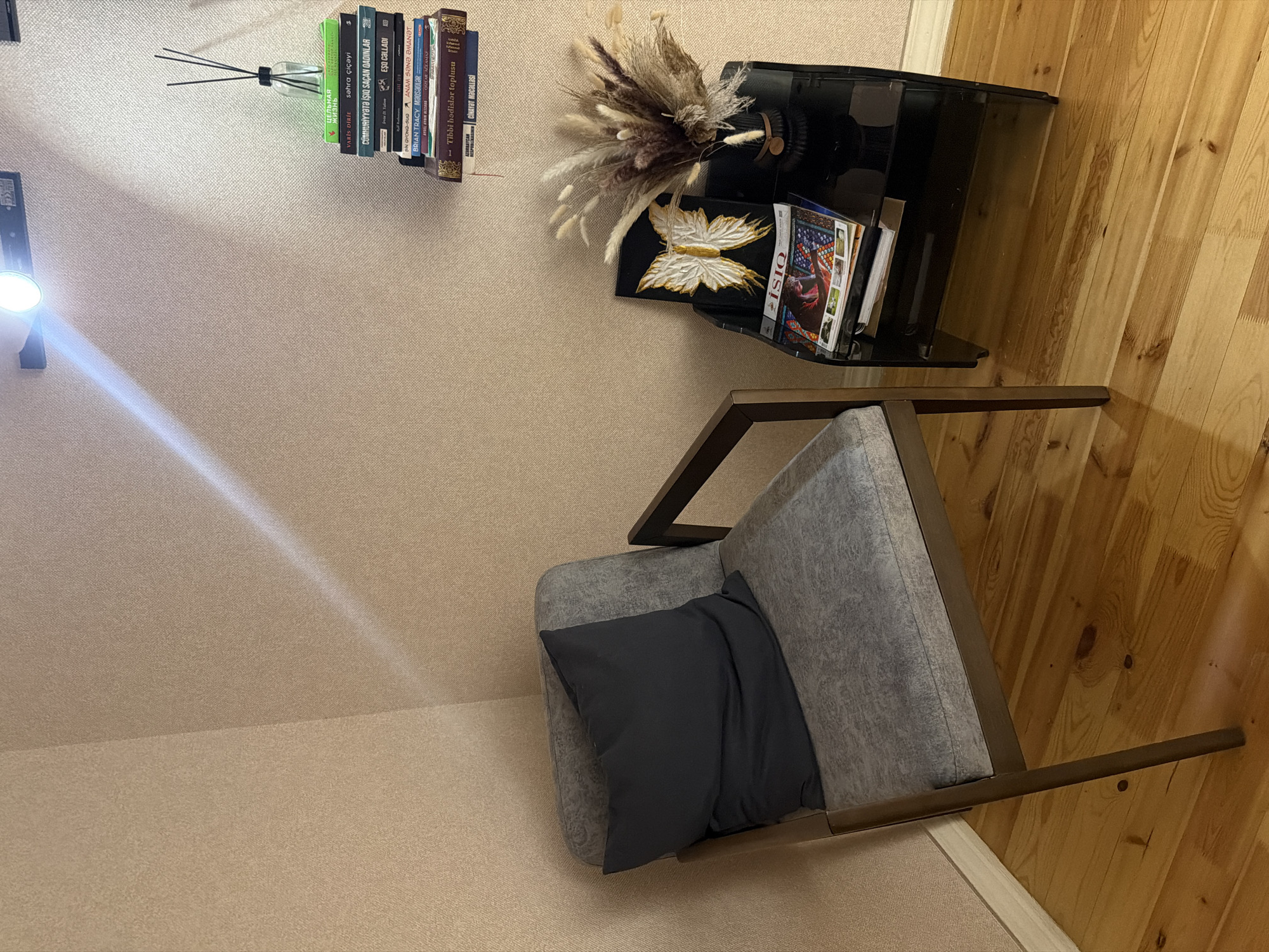 The image depicts an indoor corner with light-colored, textured wallpaper and a wooden floor. On the left, a gray upholstered armchair with a dark gray cushion is placed. To the right of the armchair, a black side table holds a decorative arrangement of dried grasses and a black vase. A framed butterfly artwork is also displayed on the side table, along with a stack of magazines, one of which is titled "İSİQ." Above the side table, a shelf is mounted on the wall, displaying a stack of books and a bottle with diffuser reeds. A light fixture is attached to the wall above the shelf, casting a bright illumination. The scene appears to be set in a room within Sabirabad, Azerbaijan, possibly a waiting area or a reading nook.