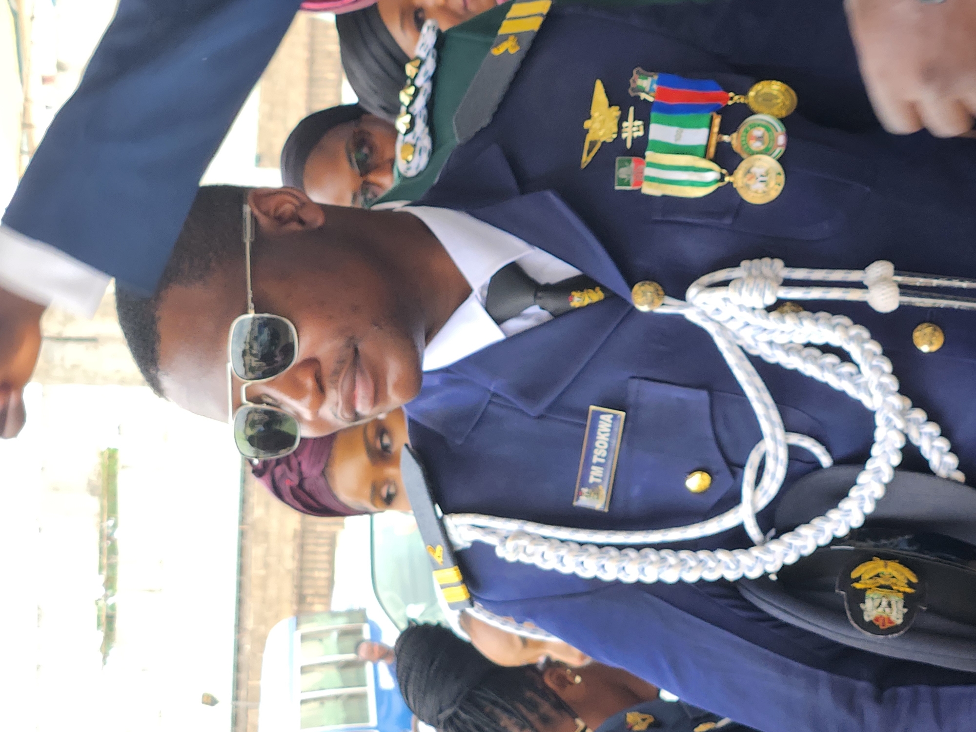 In a medium shot, a Black man in a formal navy blue uniform with white braided cords and multiple medals pinned to his chest stands in what appears to be a public outdoor setting in Barden Yana, Nigeria. He wears aviator-style sunglasses and a black tie with a small insignia. The man's name tag, "TM TSOKWA," is visible on his uniform. Behind him, several people are visible, including women wearing headscarves. A white bus with blue trim is partially visible in the background, suggesting an urban or semi-urban environment. The overall atmosphere appears to be one of formality and possibly a public event.