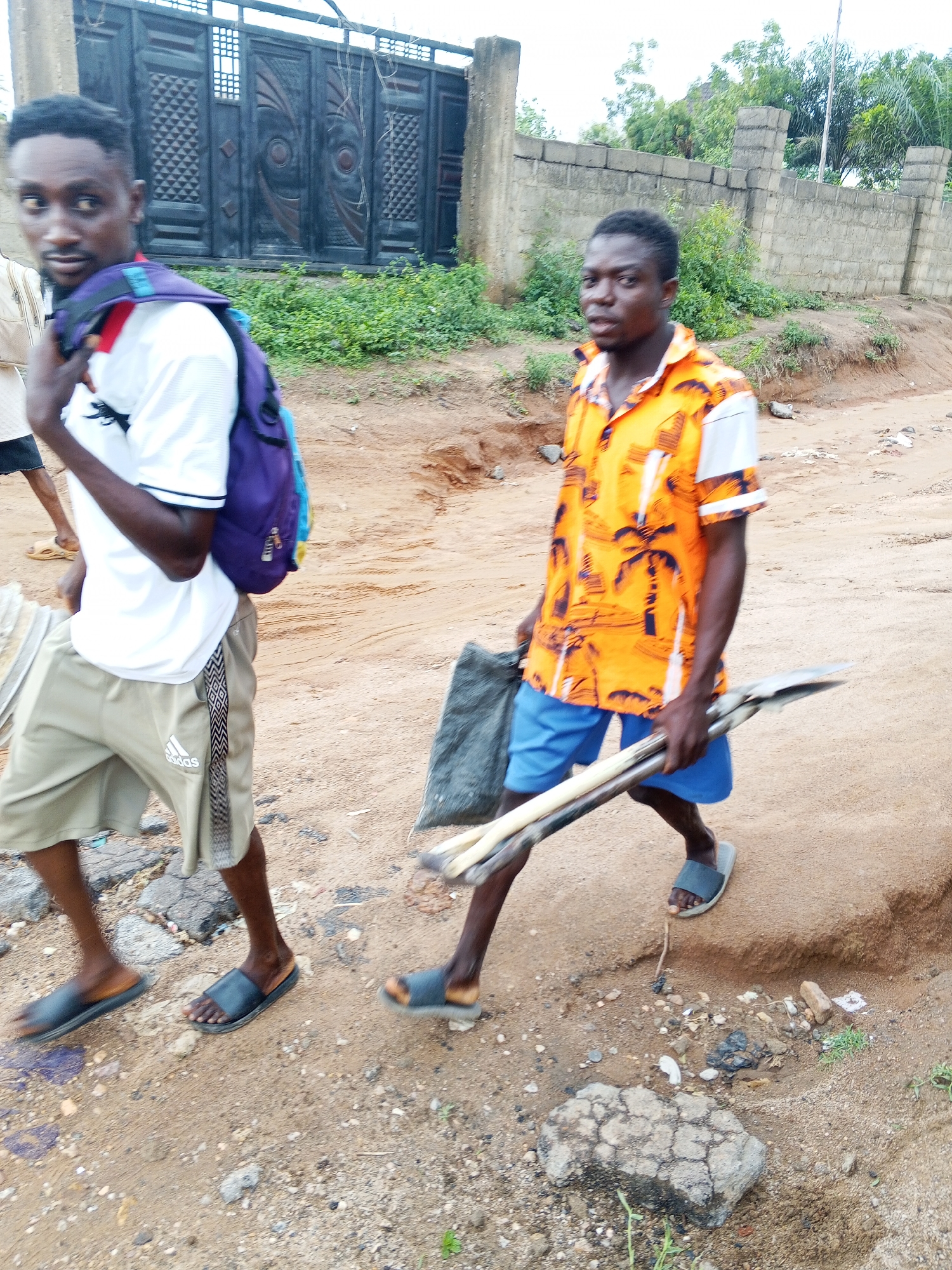 Two men are walking on a sandy, unpaved road. The man on the left wears a white shirt, khaki shorts, sandals, and a purple backpack. The man on the right wears an orange patterned shirt, blue shorts, and sandals, and carries sticks and a gray object in his hands. Behind them is a gate and a concrete wall covered with vegetation. The location is Zango-ndaji, Nigeria.
