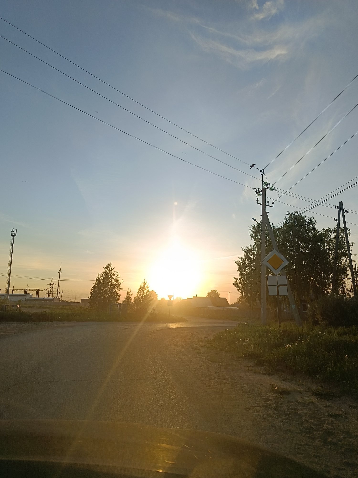 The image depicts a road at sunset near Chaikovsky, Russia. In the background are several utility poles, one with a bird perched on it and a yellow diamond-shaped road sign. There are power lines overhead. To the left, a cellular tower and a substation are visible in the distance. The sun is low on the horizon, creating a bright glare and silhouetting trees and buildings. The foreground shows the road surface, and a grassy shoulder with dandelions.