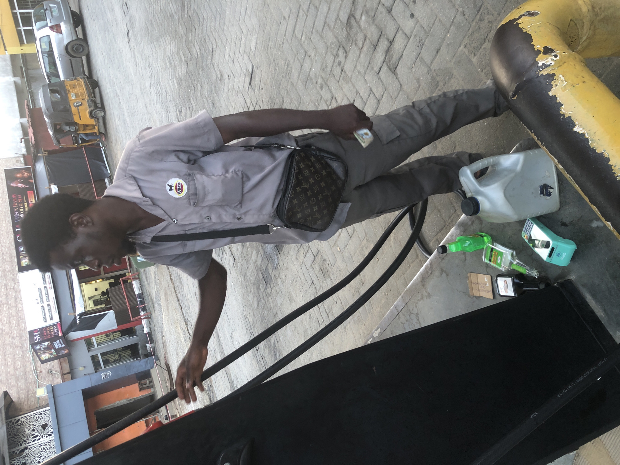 In Ado, Nigeria, a male gas station attendant, wearing a grey short-sleeved uniform shirt with a circular emblem on the left chest and grey cargo pants, is standing at a gas pump. He is holding a fuel nozzle in his right hand and is in the process of refueling a vehicle, though the vehicle itself is not fully visible. The attendant has a dark complexion and short, dark hair. He is wearing a black crossbody bag with a monogrammed pattern. In his left hand, he holds a small, rectangular item, possibly a receipt or cash.
On the ground near the gas pump are several items: a green plastic bottle, a clear plastic bottle with a white cap, a smaller brown cardboard box, a clear plastic jug with a black cap, and a light blue electronic device, possibly a credit card reader. The ground is paved with grey interlocking bricks. In the background, a yellow auto-rickshaw and a silver station wagon are visible. A building with signage, including "SM," and a salon advertised as "Unisex Beauty Parlour & Spa," is also in the background. The scene suggests a typical day at a gas station in an urban environment.