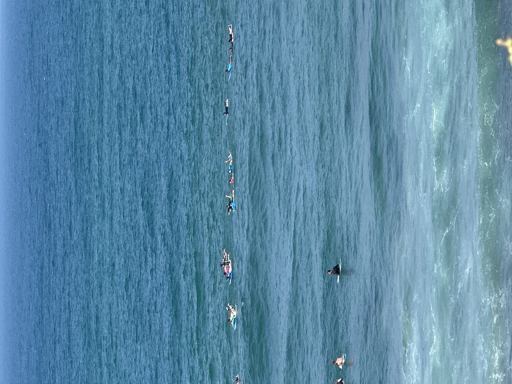 An aerial wide shot captures a group of surfers on surfboards in the clear blue ocean, near the shore of Tel Aviv, Israel. The surfers are spread out, some paddling on their boards and others sitting. The water is calm with small waves breaking closer to the shore. The sky is clear and a bright blue. The scene conveys a sense of leisure and recreation in a coastal city.