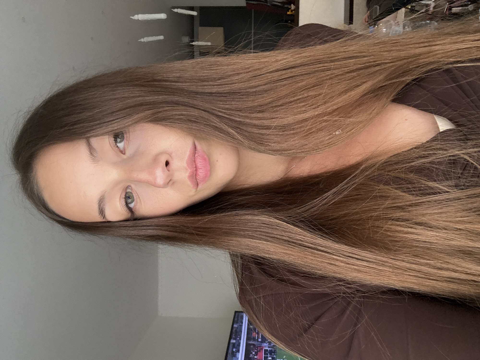 A young woman with long brown hair is facing the camera in an indoor setting. The wall behind her is grey, and there are white shapes near the ceiling. A television screen is visible below. The location in Saint Petersburg, Russia, cannot be determined from the image.