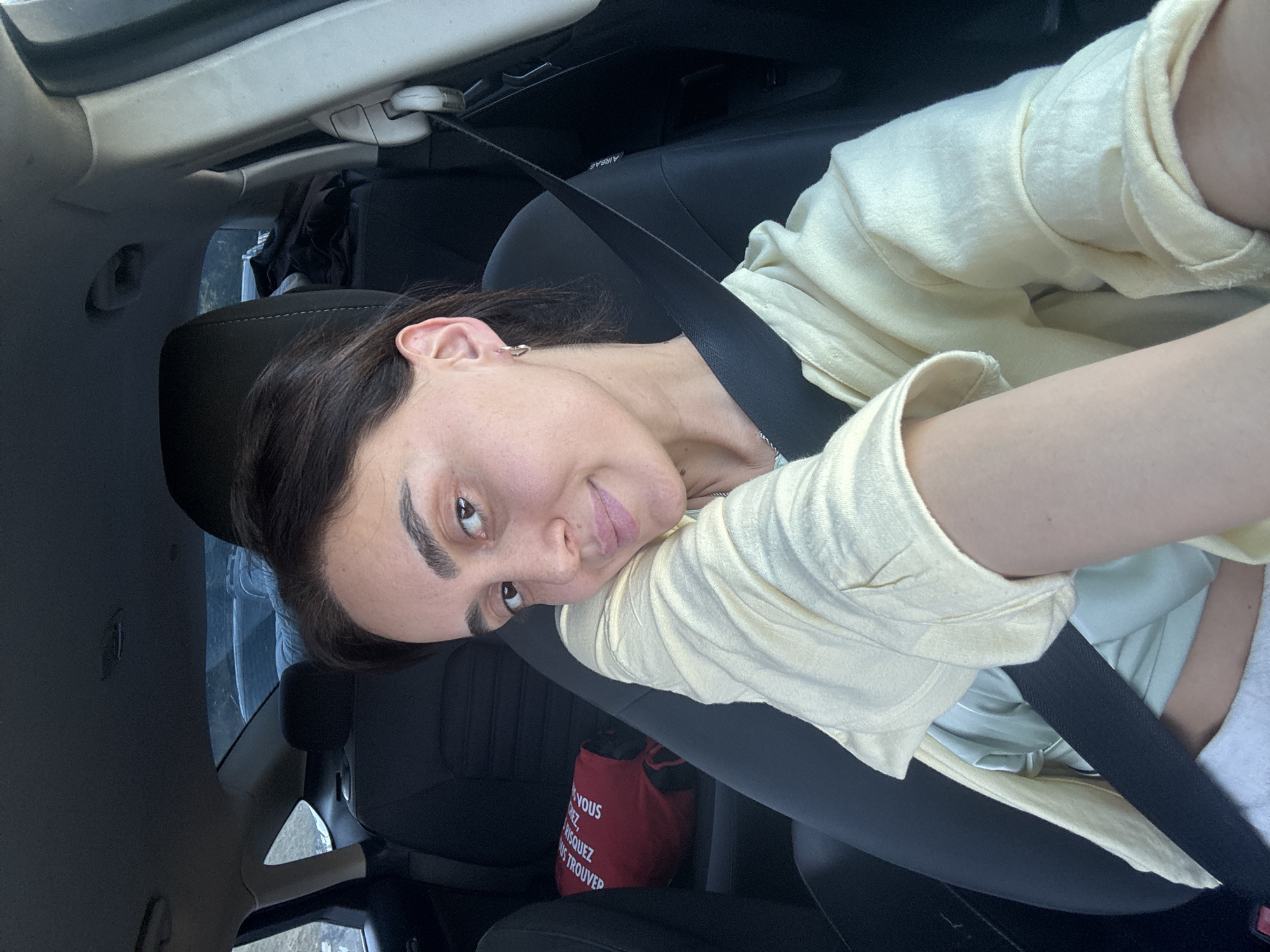 A medium shot captures a woman with dark hair, a light complexion, and prominent eyebrows, seated in the driver's seat of a car. She is wearing a light yellow collared shirt over a pale green garment and has a seatbelt fastened across her body. Her left arm is bent, resting on her shoulder, and she is looking towards the camera with a slight smile. A red bag with text in French is visible on the passenger seat behind her. The interior of the car is dark, with black seats and headrests. The windows show a blurry outdoor scene, suggesting the car is in motion or parked with a view of the surroundings. The image appears to be a selfie taken inside a car. The metadata indicates the image was taken in Tbilisi, Georgia.
