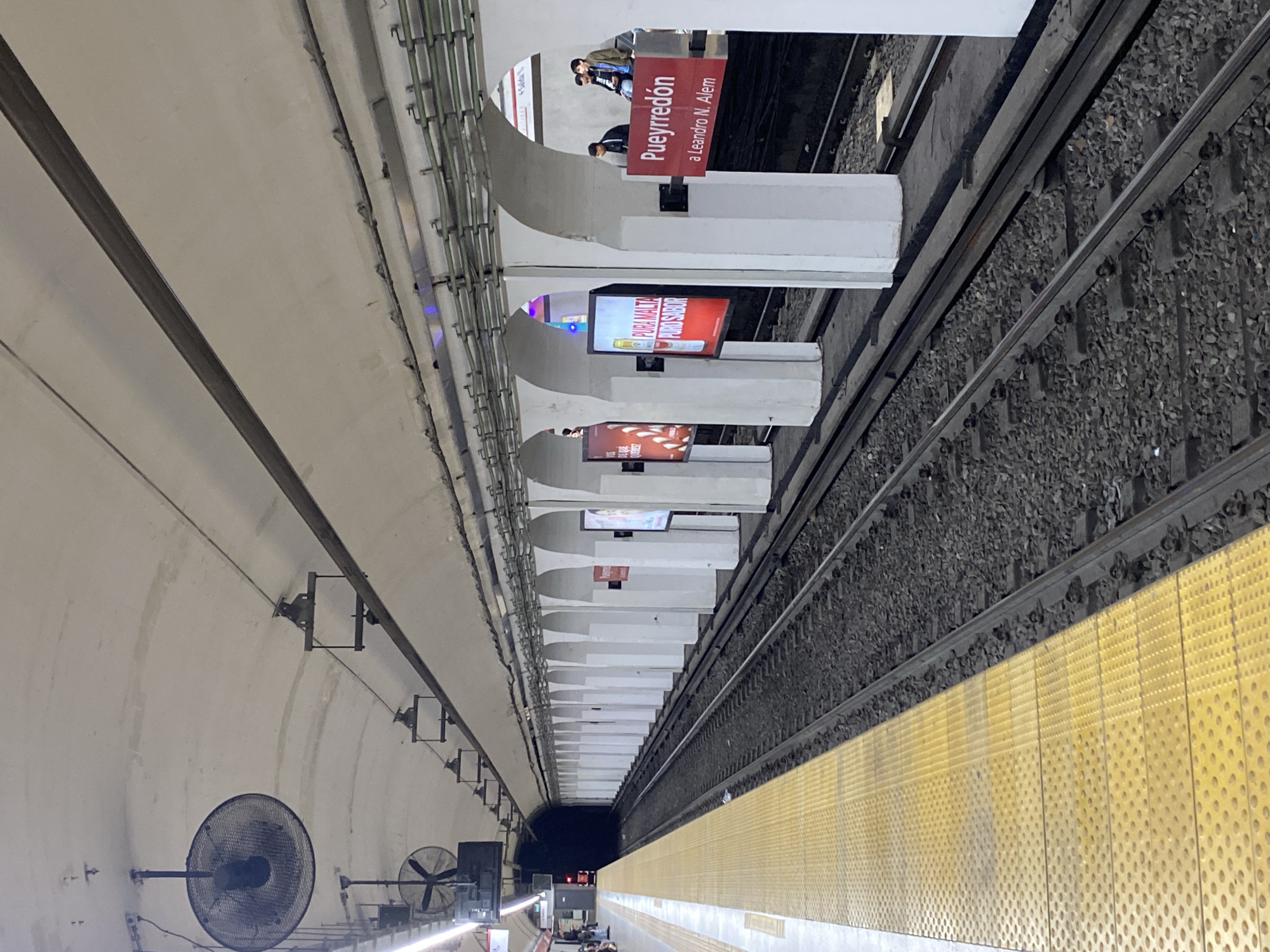 The image depicts the Pueyrredón subway station in Buenos Aires, Argentina, on Line D, heading towards Leandro N. Alem. The station features a distinctive architectural design with arched supports along the platform. Tracks run adjacent to the platform, filled with gravel. A yellow tactile paving strip runs along the platform edge. Overhead, there are fans, and advertisements are visible on the station signage. People can be seen waiting on the platform in the direction of Leandro N. Alem.