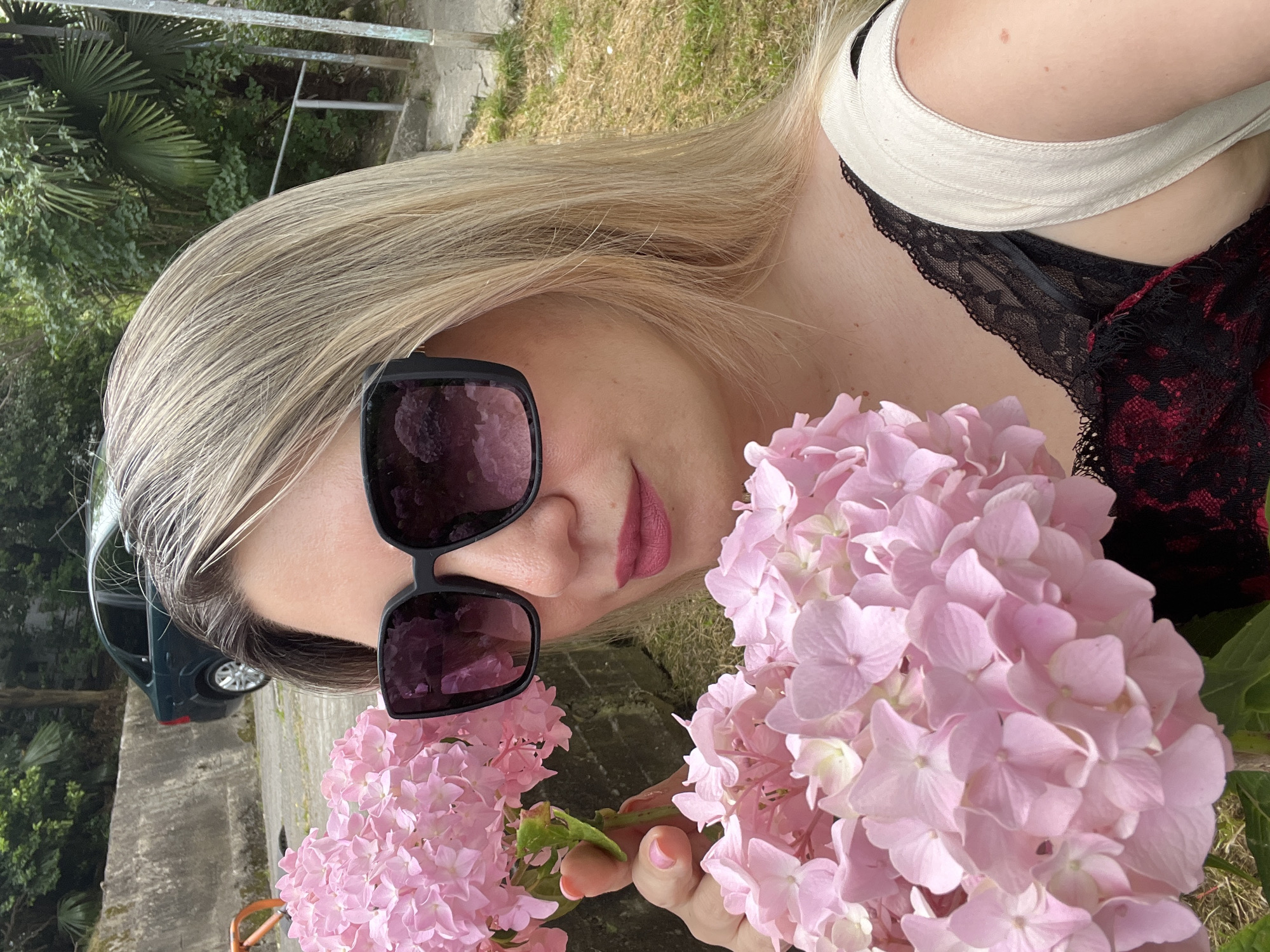 A medium shot captures a blonde woman with fair skin and medium complexion in a self-portrait, holding a bouquet of pink hydrangeas. She is wearing large, dark-rimmed sunglasses, and her lips are painted with pink lipstick. Her hair is blonde with lighter streaks, falling past her shoulders. She is wearing a black lace top beneath a beige garment. The background features lush green foliage, a concrete wall, and a dark green sedan parked on a paved area. The scene appears to be outdoors, possibly in a park or garden setting within the city of Sochi, Russia, indicated by the presence of palm trees and other vegetation typical of the region. The overall mood is relaxed and summery.