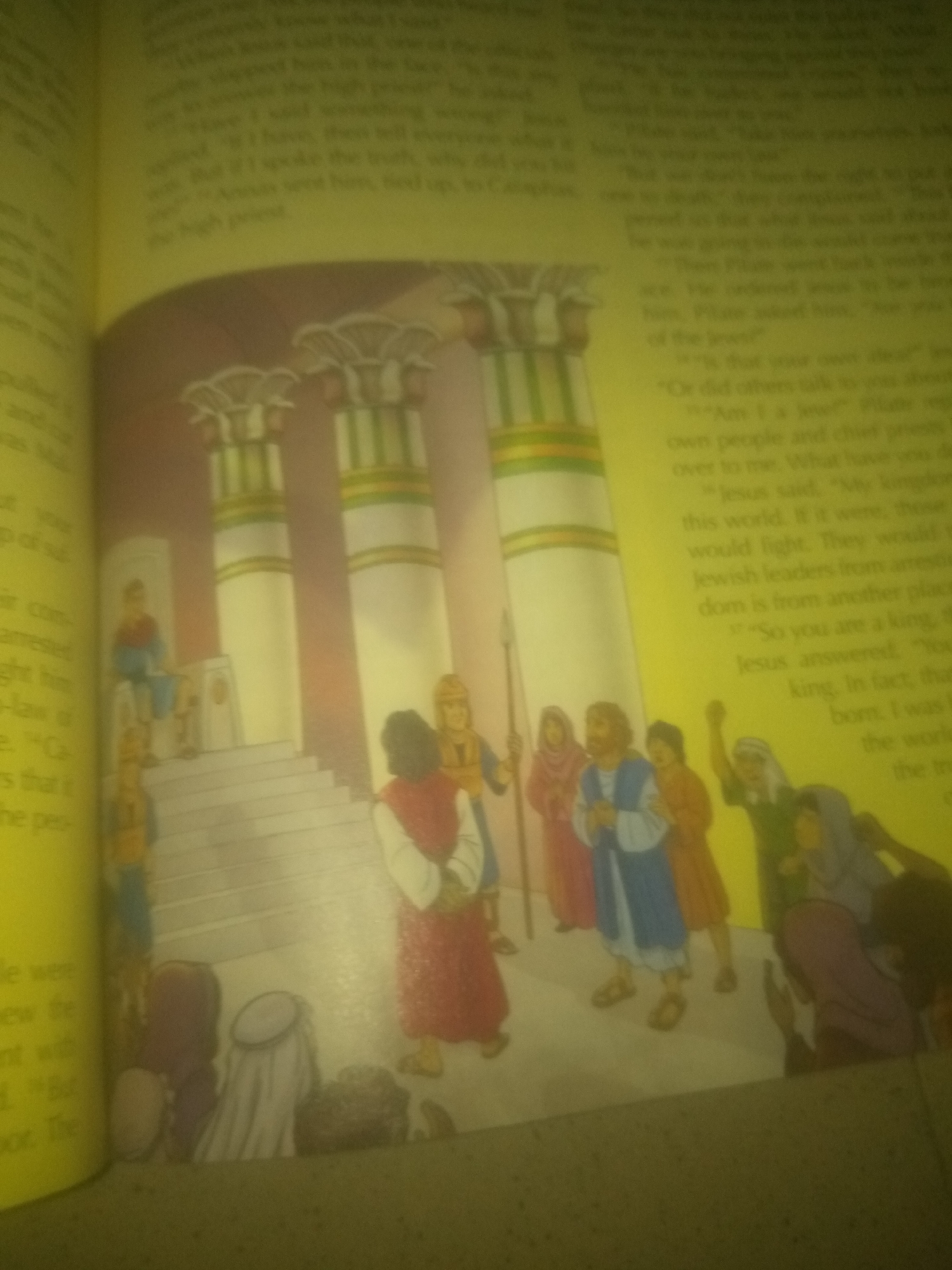 The image shows an illustration from a book depicting a scene with several people and architectural elements. On the left side of the image, there is a figure seated on a throne-like structure, with stairs leading up to it. This figure appears to be dressed in blue and red. In front of the throne, there are two figures standing, one in red and white, and another in what appears to be golden armor with a red cap. To the right of these figures, a group of people are gathered, some in colorful robes. One individual in a blue and white robe stands out. Behind this group, there are large, fluted columns with decorative tops. The background appears to be a pale yellow, suggesting an indoor or stylized setting. The text on the left and right sides of the illustration contains what appears to be a biblical narrative. There is no information or visual context within the image to suggest a location in Jauro Yinu, Nigeria.