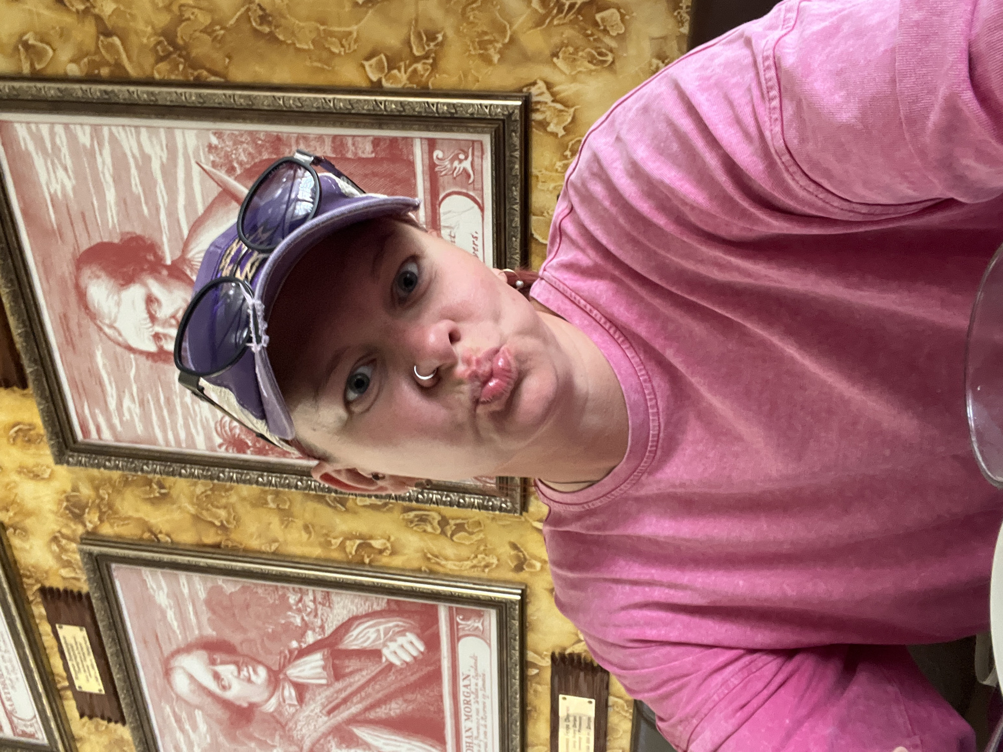 A selfie of a fair-skinned person with short hair, wearing a pink t-shirt, a purple baseball cap, and sunglasses on their hat. They have a nose ring and are pursing their lips in a kissing expression. The background features framed portraits hanging on a patterned gold wall. Text on the bottom portrait reads "JOHAN MORGAN." The location is identified as Chaykovsky, Russia.