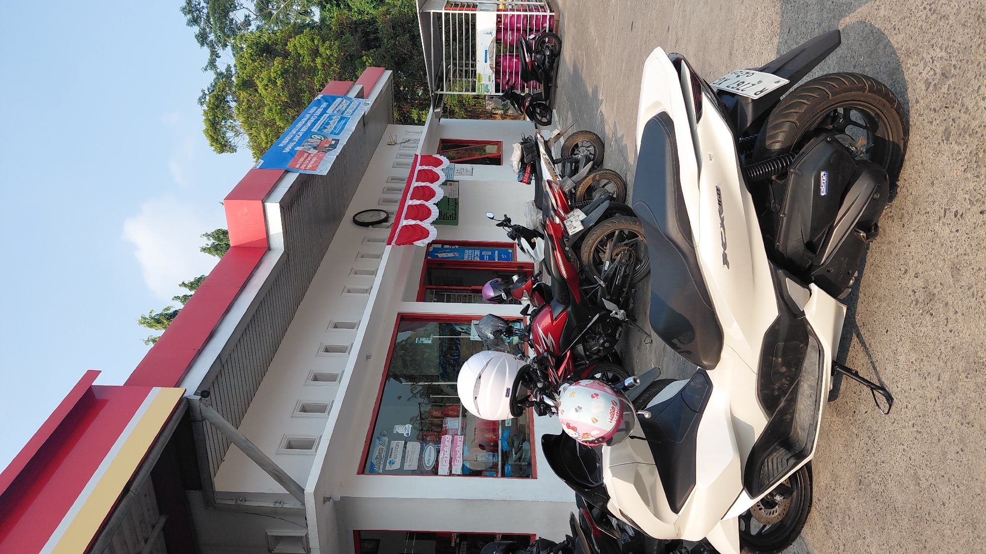 The image captures a view of a gas station in Karanglewas, Indonesia, with multiple motorcycles parked in front of a building. The building features a white facade with red trim along the roofline and an awning with red and white scalloped fabric above the entrance. A sign with blue text and images is attached to the upper portion of the building. Several motorcycles are visible, including a prominent white scooter in the foreground, a red motorcycle with a white helmet on its handlebars, and other motorcycles parked further back. One motorcycle has a helmet adorned with a Hello Kitty design. The ground is paved with concrete. Trees and foliage are visible in the background. A clock is mounted on the building's exterior. The sky is clear with some light clouds.