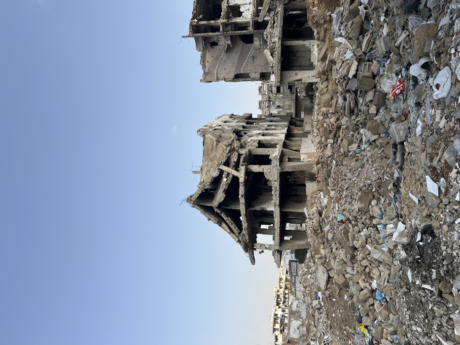 This image depicts a scene of extensive urban destruction in Joubar Municipality, Syria. The foreground is dominated by a vast expanse of rubble composed of broken concrete, bricks, rebar, and miscellaneous debris, including scattered plastic bags and wrappers.
In the midground, two severely damaged multi-story concrete buildings are prominent. The building on the left features a partially collapsed roof and upper floors, forming a jagged, angular silhouette with exposed rebar and what appear to be remnants of antennae on its remaining structure. Its lower floors show large gaping openings where walls and windows once stood. To the right, another multi-story concrete structure exhibits significant damage, with large sections of its facade missing, revealing an internal staircase with visible steps and railings. Its exterior walls display numerous small circular impacts consistent with bullet holes.
The background extends into a panorama of further damaged and partially collapsed buildings and mounds of debris under a clear, light blue sky with a faint, wispy cloud. No people, animals, or active interactions are visible. The environment is characterized by widespread destruction, dust, and sparse, dry vegetation near the base of the left building.