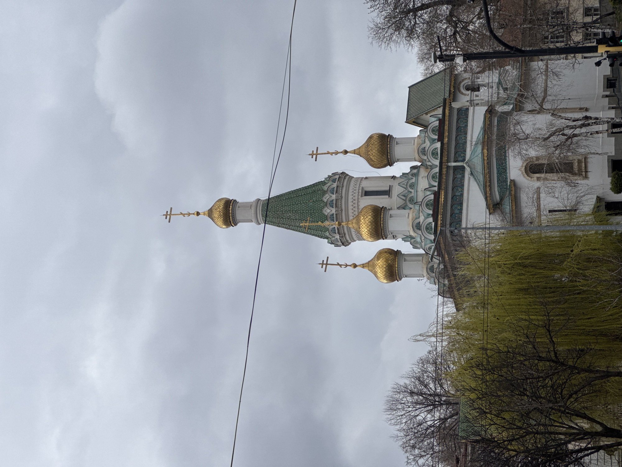 The image captures the Russian Church of Saint Nicholas the Miracle-Maker in Sofia, Bulgaria. The architectural style is distinctively Russian Revival with green tiled roofs and golden onion domes topped with Orthodox crosses. The church is white with ornate decorations. A power line runs across the cloudy sky in the background. Bare trees and one willow tree are in the foreground, indicating an early spring or late autumn setting. There are also traffic signals present, showing the setting is the city, and other buildings around it.
