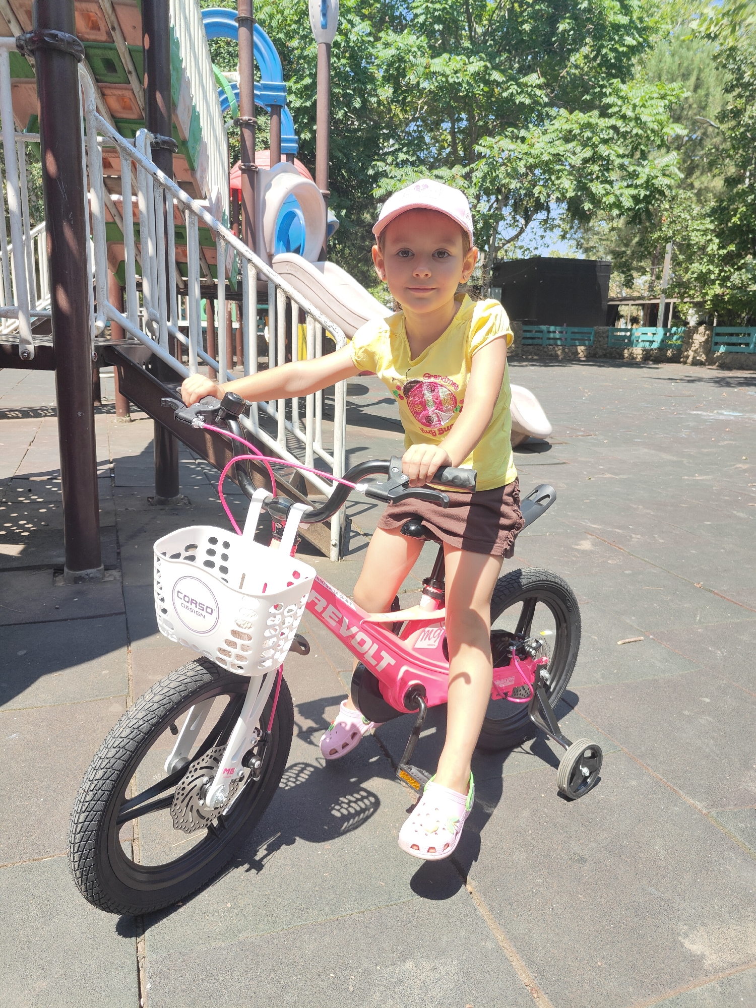 A young girl with blonde hair, wearing a yellow t-shirt and a pink baseball cap, sits on a pink children's bicycle in a park. The bicycle has a white basket on the front with the "CORSO DESIGN" logo. The girl is wearing pink Crocs. In the background, there is a playground structure with slides and stairs, and trees. The ground is paved with dark grey rubber tiles. The scene appears to be a sunny day. The location is identified as a park in Chornomorsk, Ukraine.