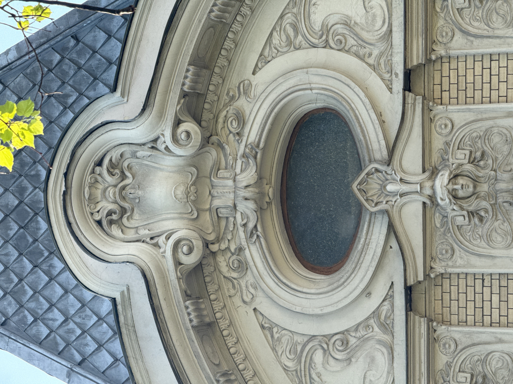 The image shows an ornate, vertically oriented architectural detail of a building facade. The architectural style appears to be from the Beaux-Arts period. The details consist of scrolling acanthus leaves, rosettes, and classical motifs. The left side of the frame features a section of a dark, patterned surface with square tiles, likely roofing or cladding. Adjacent to this is a sculpted, light-colored architectural element with complex moldings and scrollwork. A central oval opening filled with a dark material is visible. To the right, there are brickwork patterns and further embellishments, including a sculpted face. The building is located in an unknown city or state.