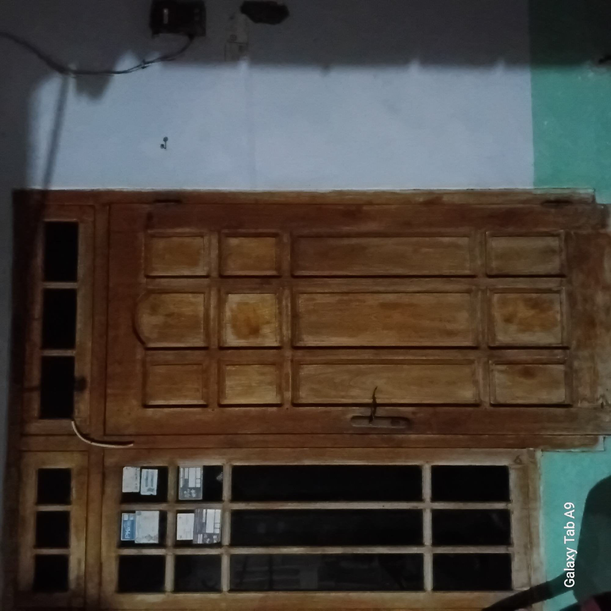This is a medium-shot, eye-level, interior photograph of a wooden door and adjacent window with a plain wall to the right. The door is made of dark, aged wood with a grid pattern of square panels, some of which have a lighter, stained appearance. A simple metal door handle is visible on the left side of the door. The window to the left of the door is also made of wood and has a grid of six vertical panes and two horizontal panes at the top. Some papers are affixed to the panes of the window. The wall to the right is painted a light color and shows some staining and shadows. A dark electrical box is mounted on the wall near the top right corner. The bottom of the wall is painted a greenish color. In the bottom left corner of the image, the text "Galaxy Tab A9" is visible. This appears to be a default watermark from a device. The overall lighting is dim, suggesting it is either evening or the area is poorly lit. The scene is likely a residential or commercial entryway in Trenggalek, Indonesia, given the mention of the device.